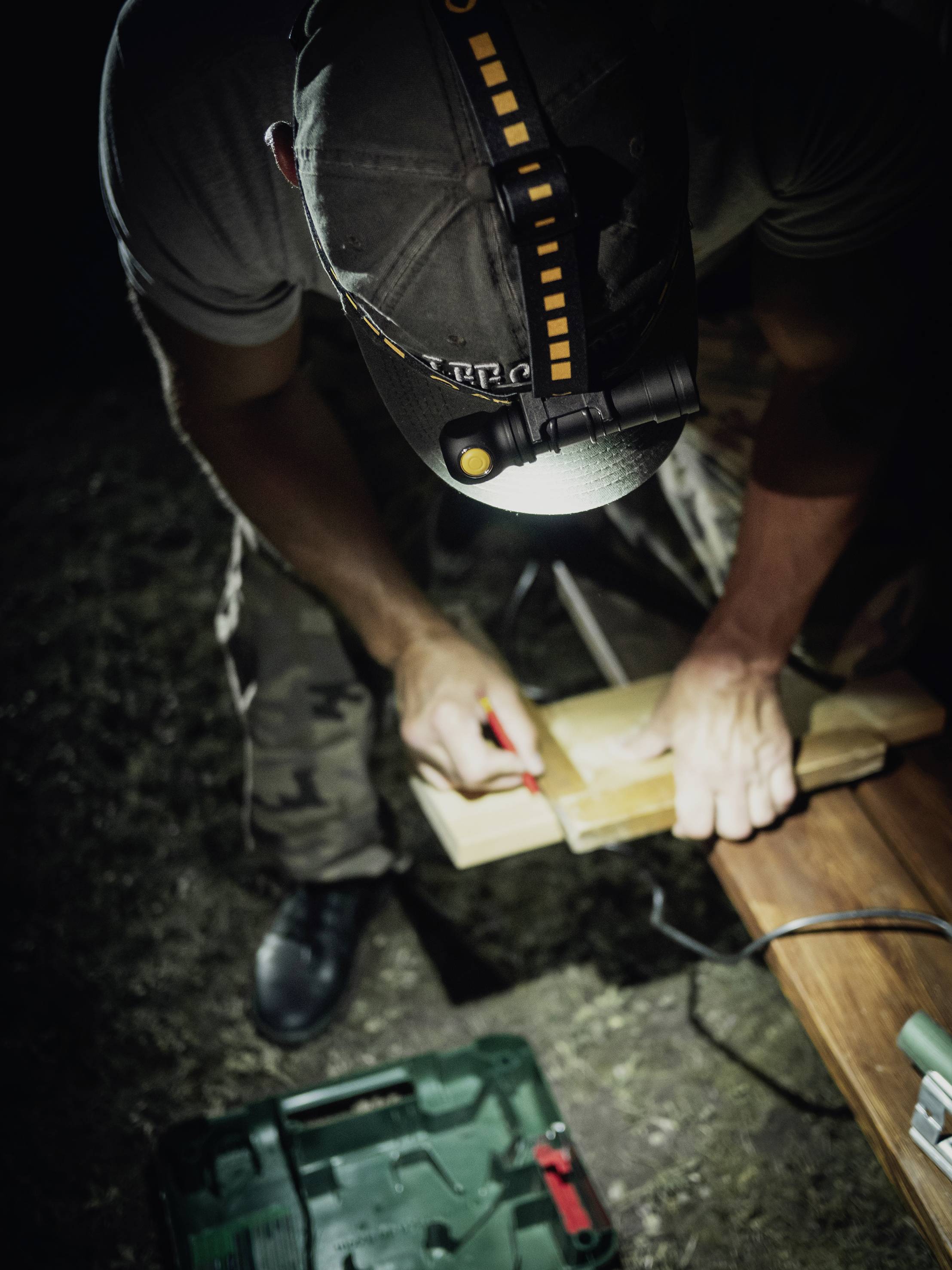 Eine Person arbeitet nachts im Freien mit einer Stirnlampe an einem Holzbrett. Vor ihnen liegt ein offener Werkzeugkasten auf dem Gras.