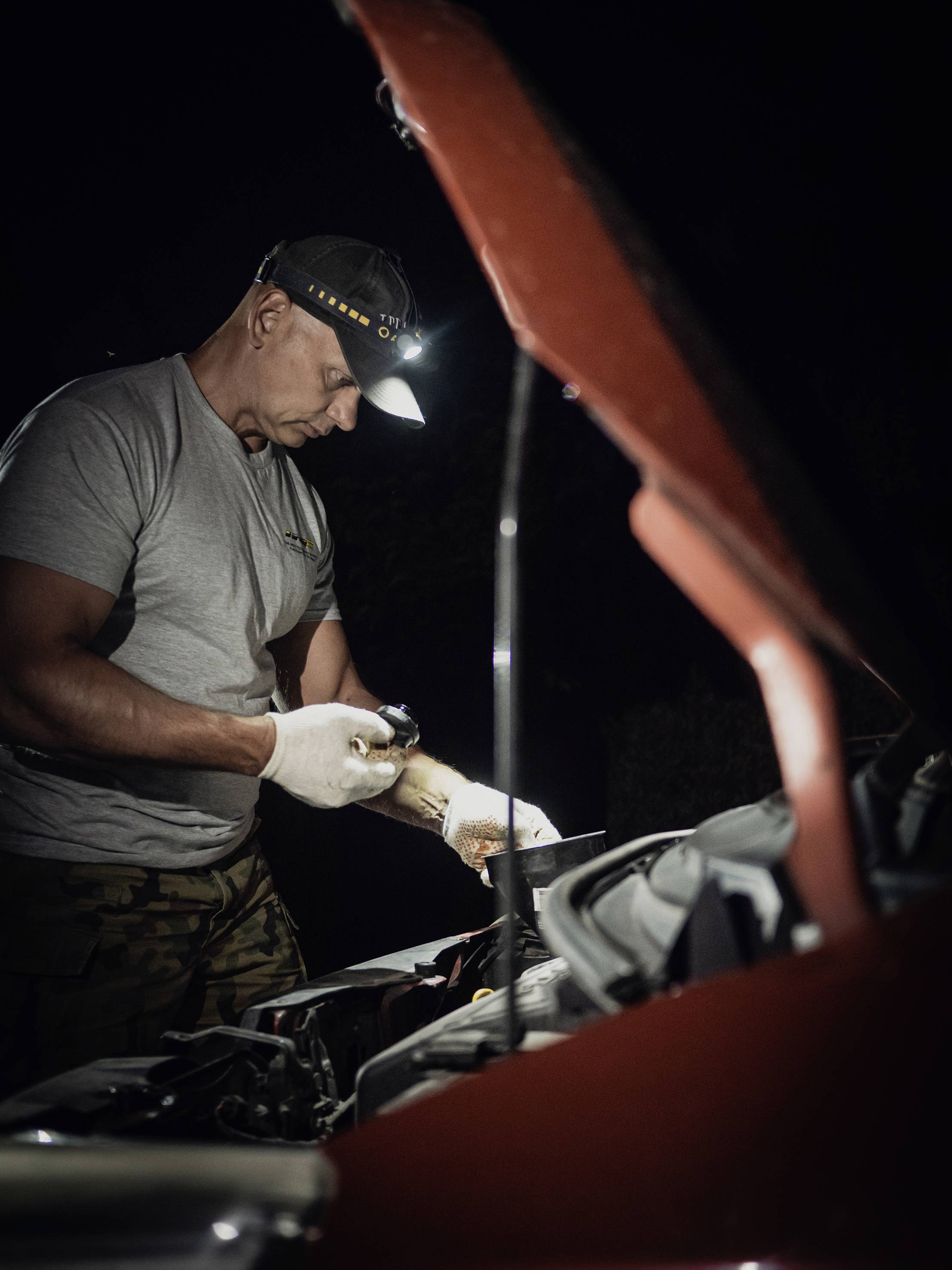 Ein Mann in grauem T-Shirt repariert nachts mit Stirnlampe Motor eines Autos mit offenem Motorhaube.