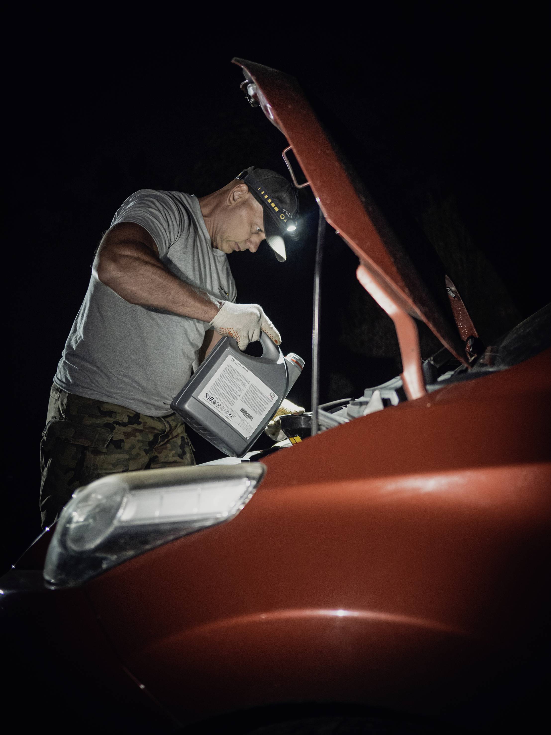 Eine Person in Arbeitskleidung fügt Öl in den Motor eines roten Autos ein. Die Szene findet nachts statt, beleuchtet von einer Stirnlampe.