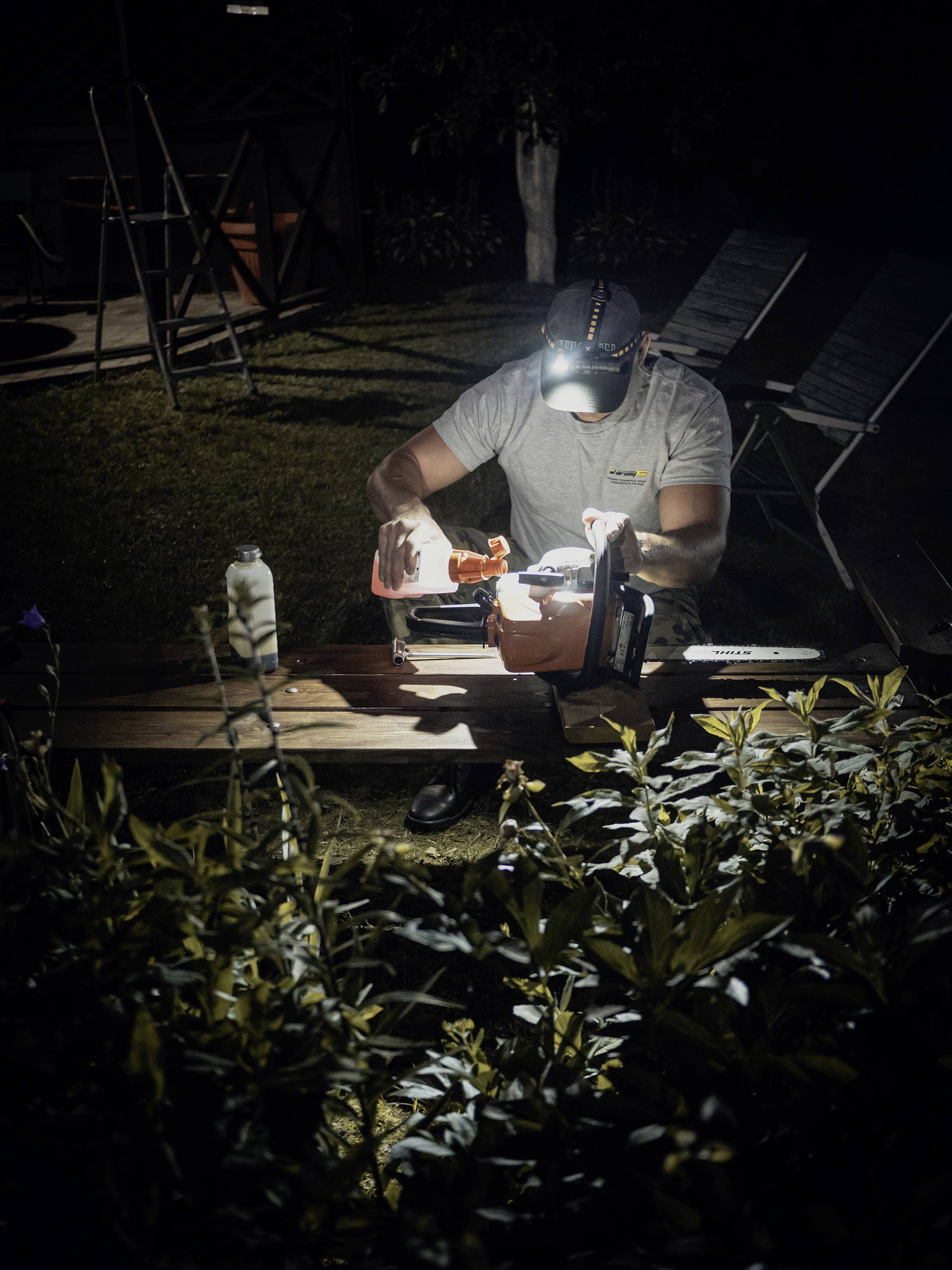 Eine Person mit Stirnlampe arbeitet nachts draußen an einem Tisch, umgeben von Pflanzen. Sie konzentriert sich auf ein Projekt.