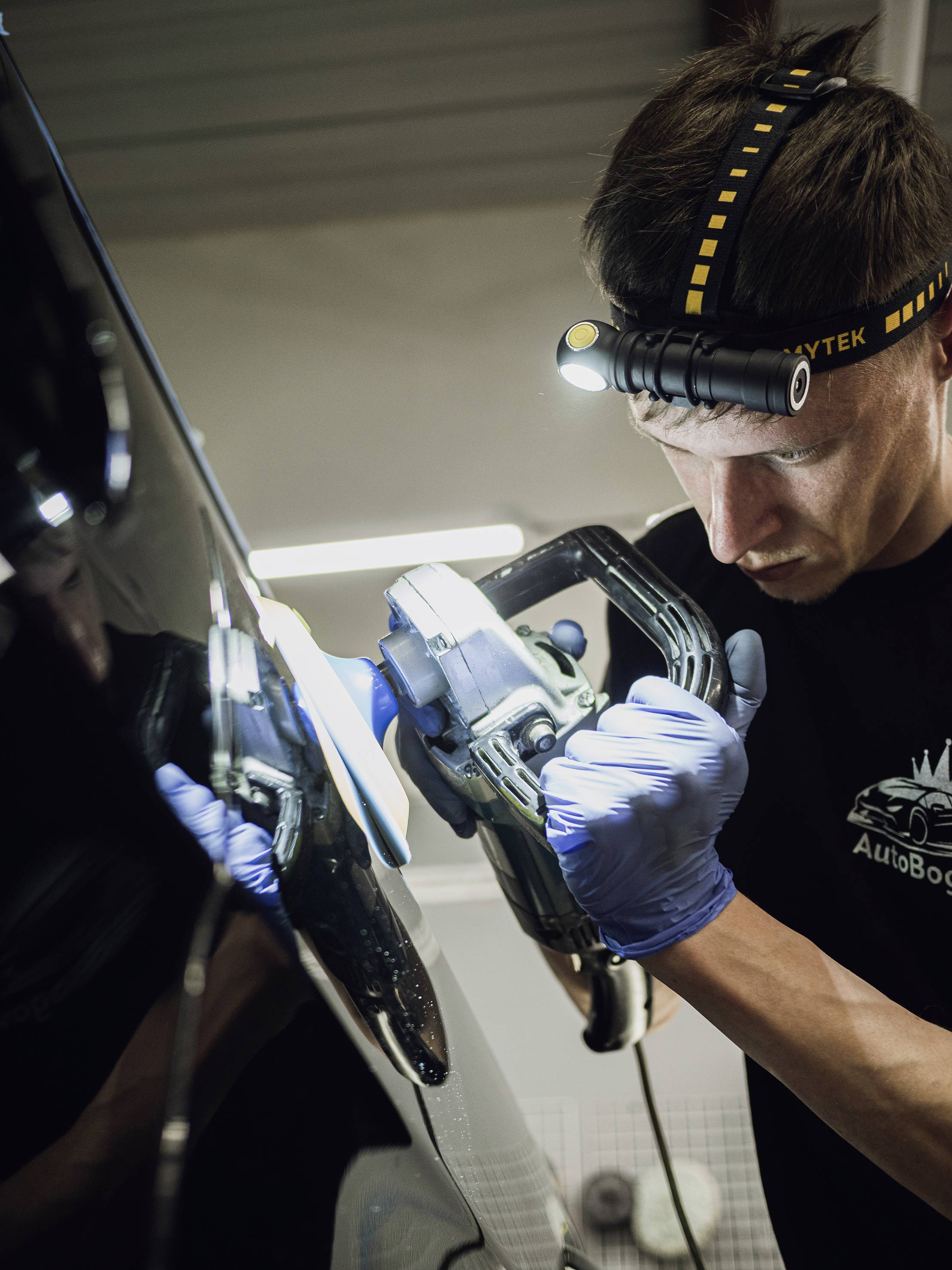 Ein Mechaniker poliert ein Auto mit einer Schleifmaschine in einer Werkstatt. Er trägt Handschuhe und eine Stirnlampe.