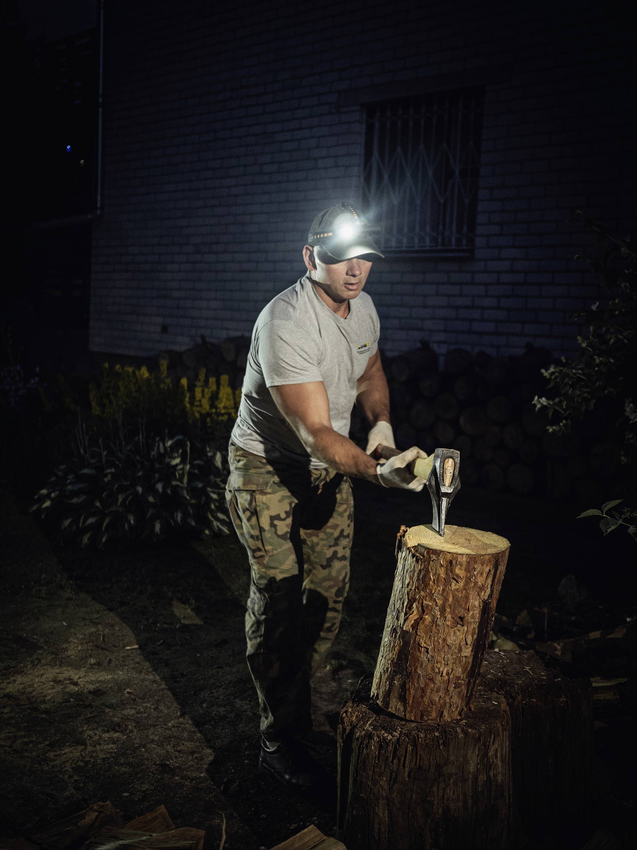 Ein Mann in Tarnhose spaltet im Dunkeln Holz in einem Garten. Er trägt eine Stirnlampe und verwendet eine Axt.