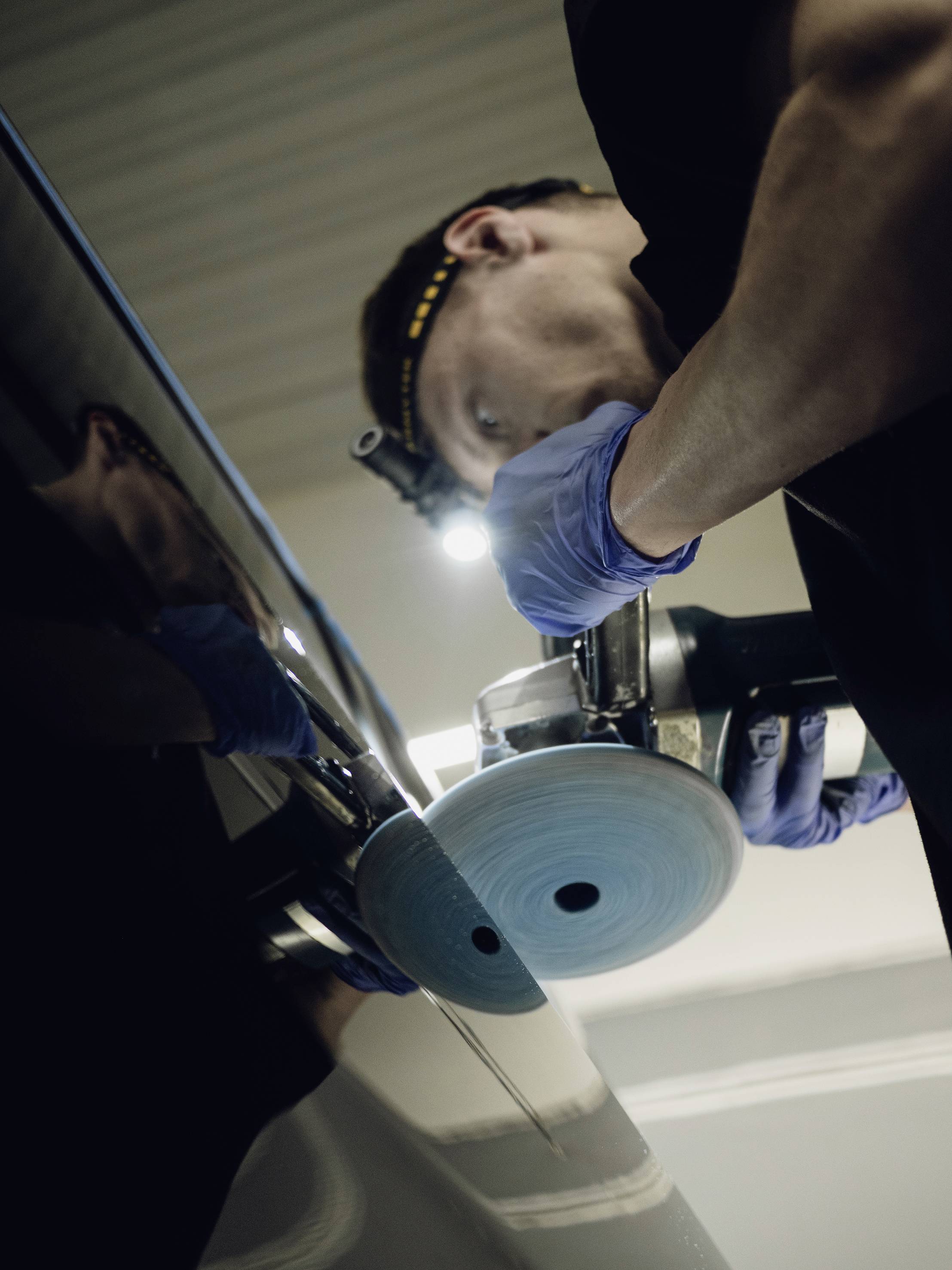 Ein Mechaniker poliert mit einem Poliergerät einen Autolack. Er trägt blaue Handschuhe und eine Stirnlampe für bessere Sicht.