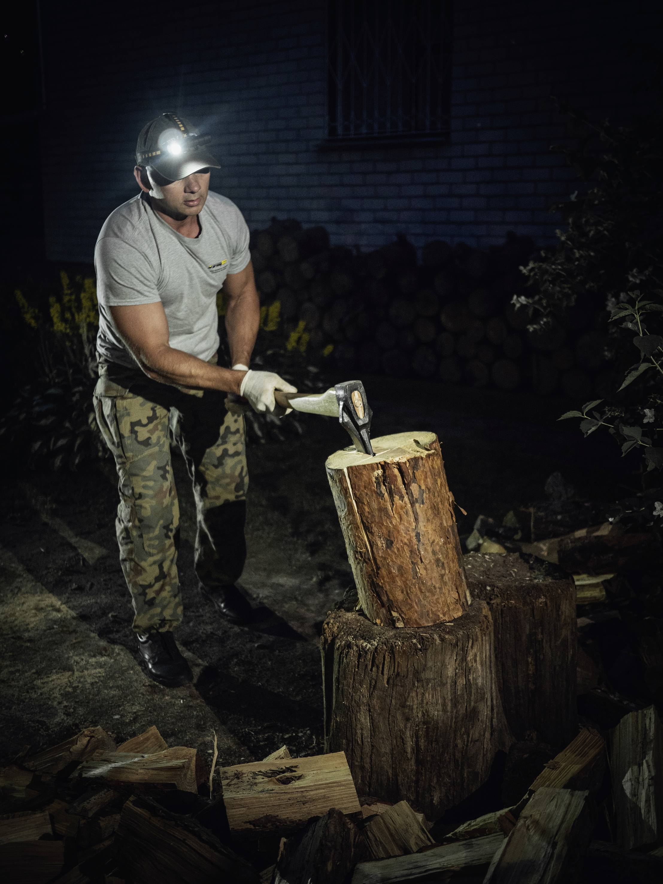 Eine Person spaltet nachts mit einer Axt Holz. Sie trägt eine Kopflampe und Tarnhose. Im Hintergrund sind Holzstapel zu sehen.