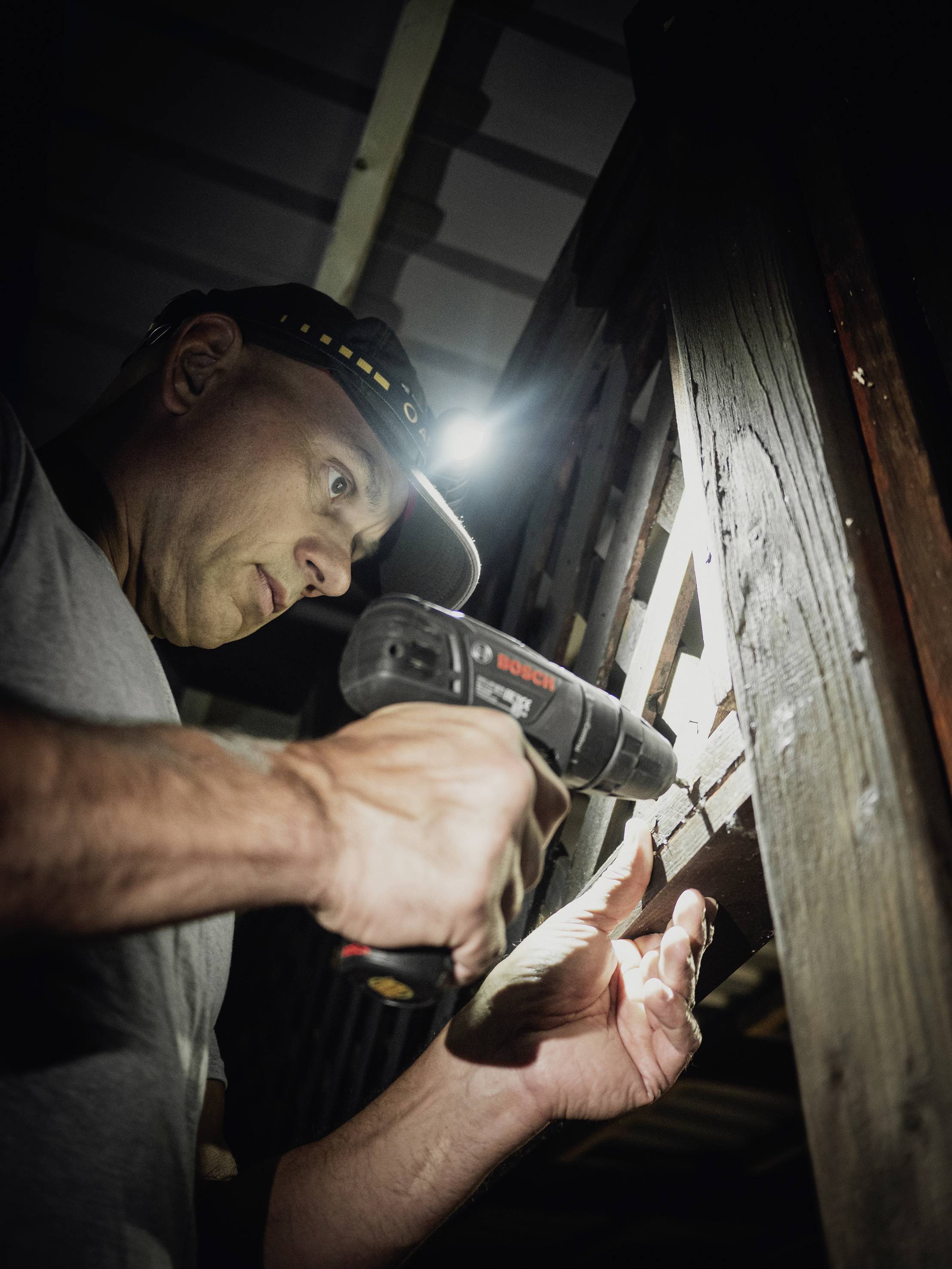 Ein Mann mit Stirnlampe benutzt einen Akkuschrauber, um Holz zu bearbeiten. Umgebungslicht ist schwach, Fokus auf der handwerklichen Arbeit.