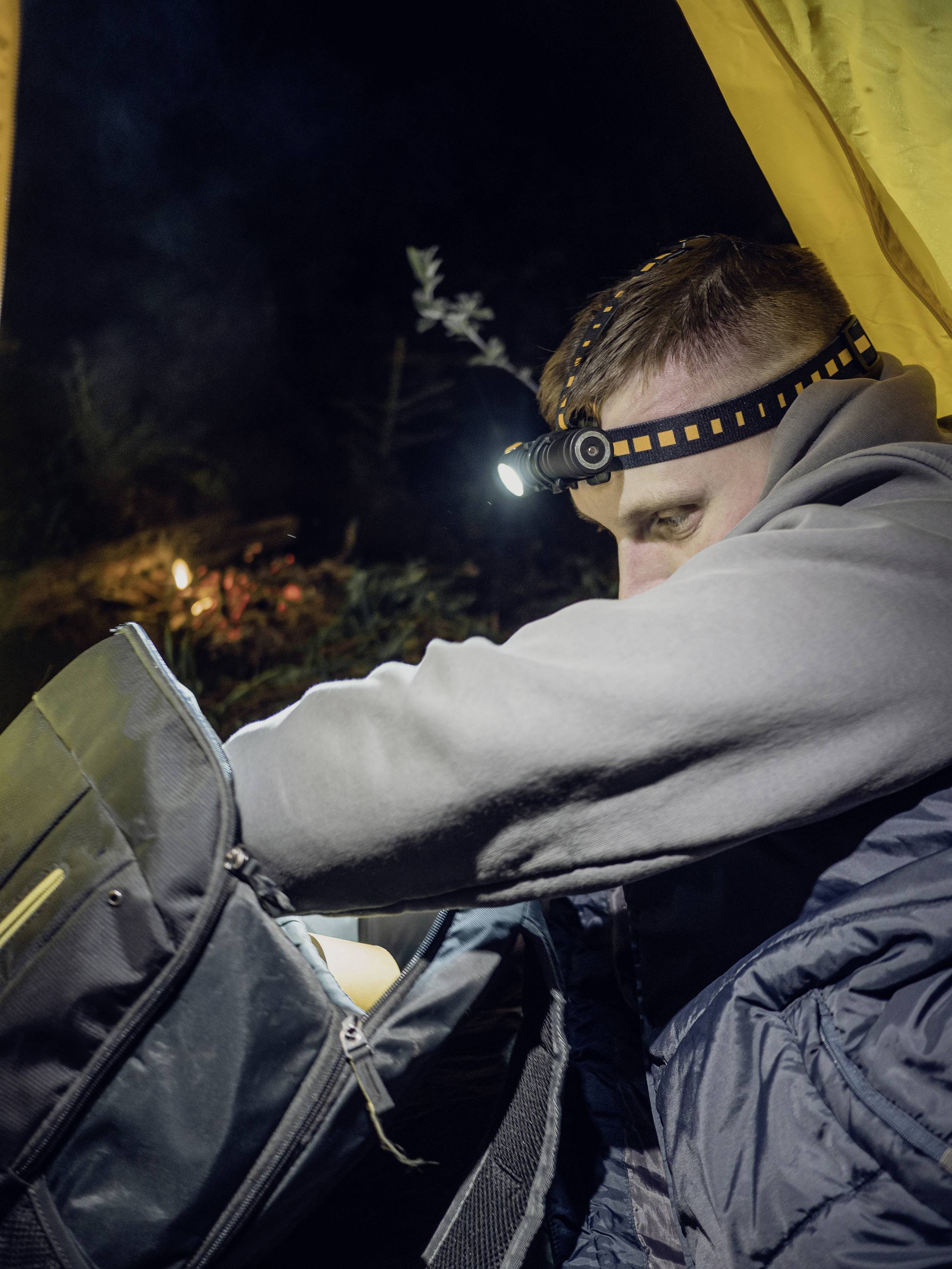 Mann in Zelt bei Nacht, trägt Stirnlampe, durchsucht Rucksack. Im Hintergrund schwaches Lagerfeuerlicht sichtbar.