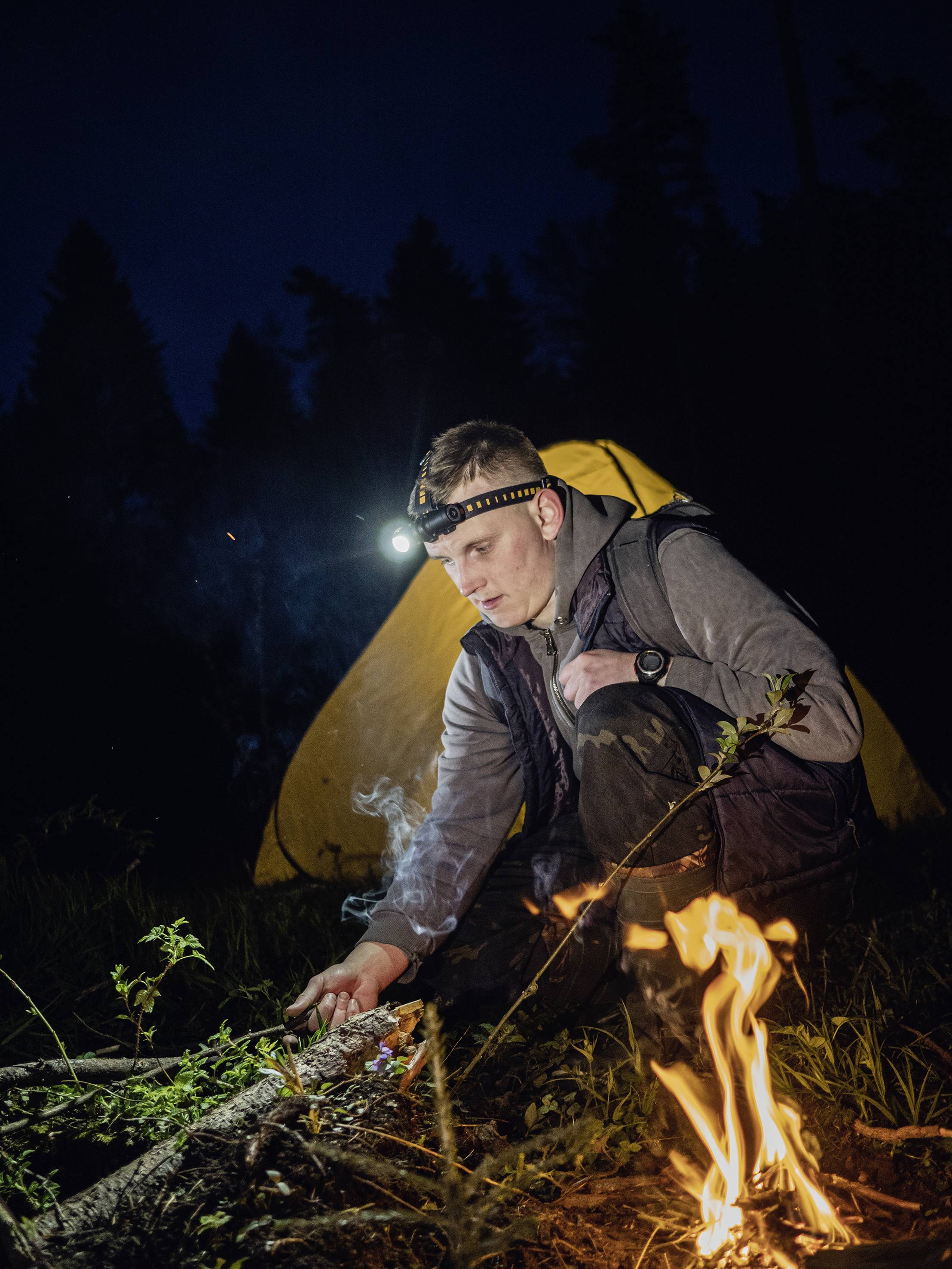 Ein Mann zündet im Schein einer Stirnlampe ein kleines Lagerfeuer an, neben einem gelben Zelt in einem bewaldeten Gebiet bei Nacht.