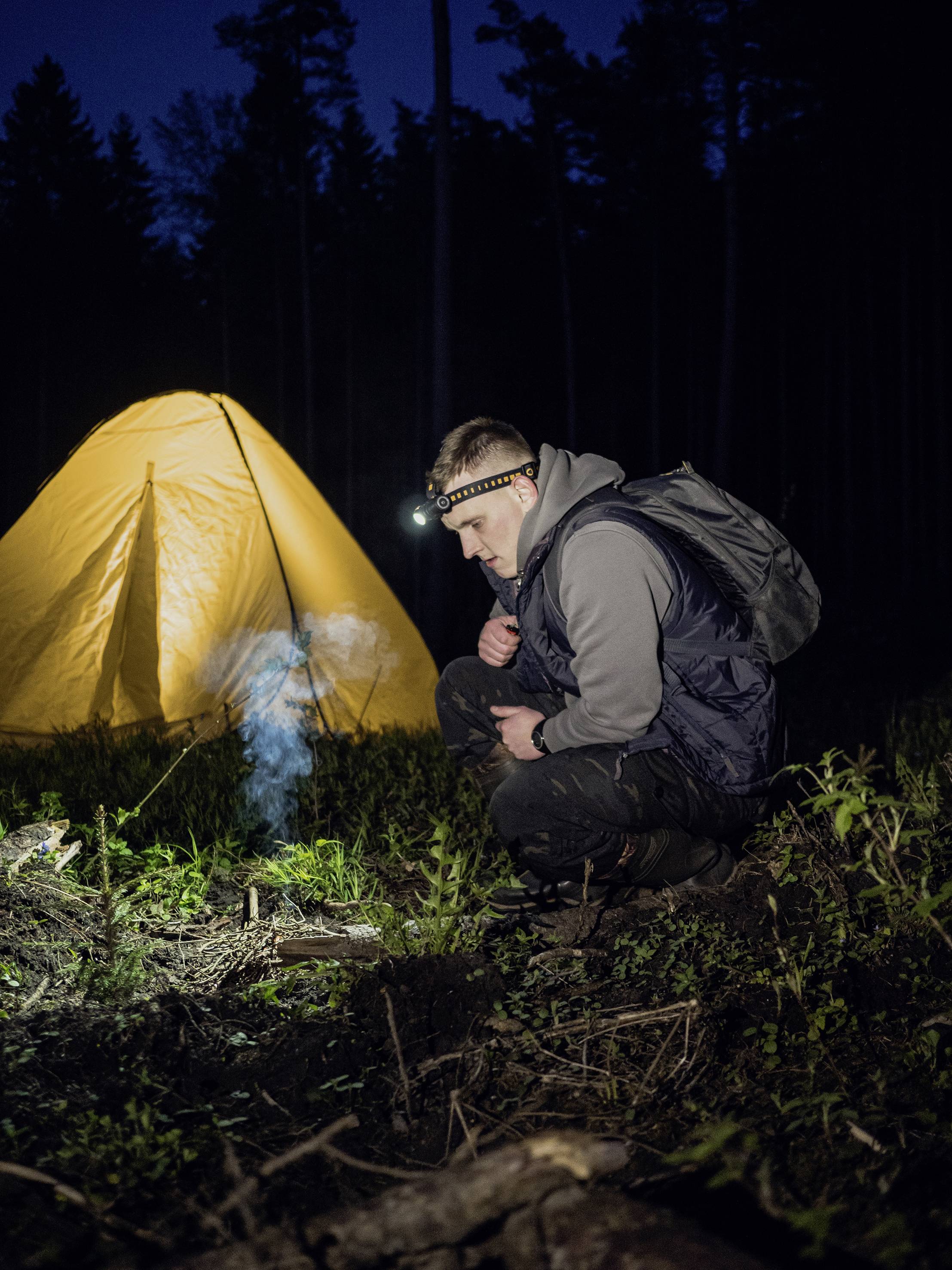 Mann kniet vor kleinem Lagerfeuer im Wald. Im Hintergrund steht ein beleuchtetes Zelt.