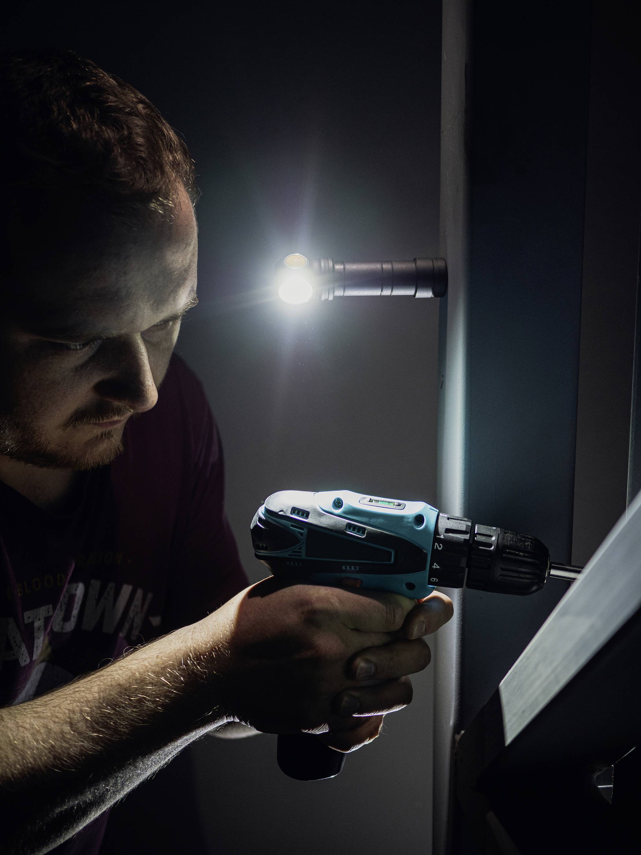 Ein Mann benutzt eine elektrische Bohrmaschine, um eine Schraube in eine Wand zu schrauben. Eine Lampe leuchtet den Arbeitsbereich aus.