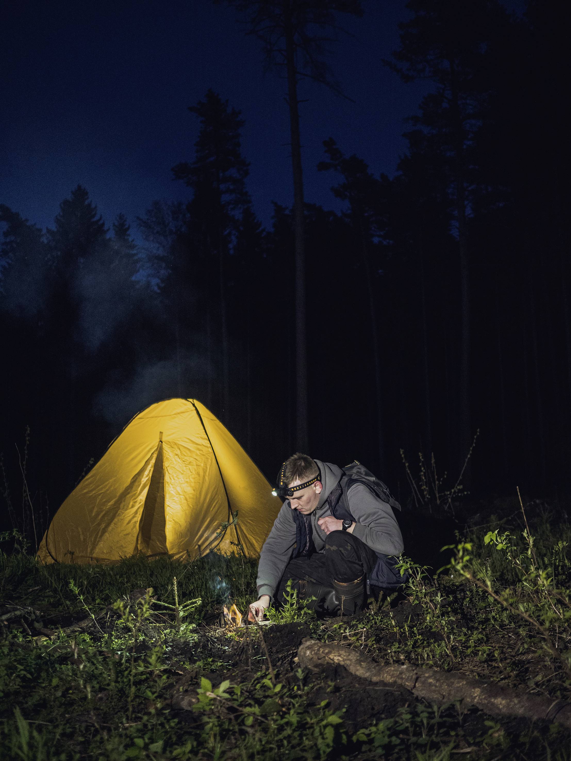 Eine Person zündet ein Feuer vor einem gelben Zelt in einem dunklen Wald an. Die Szene vermittelt Camping in der Wildnis bei Nacht.