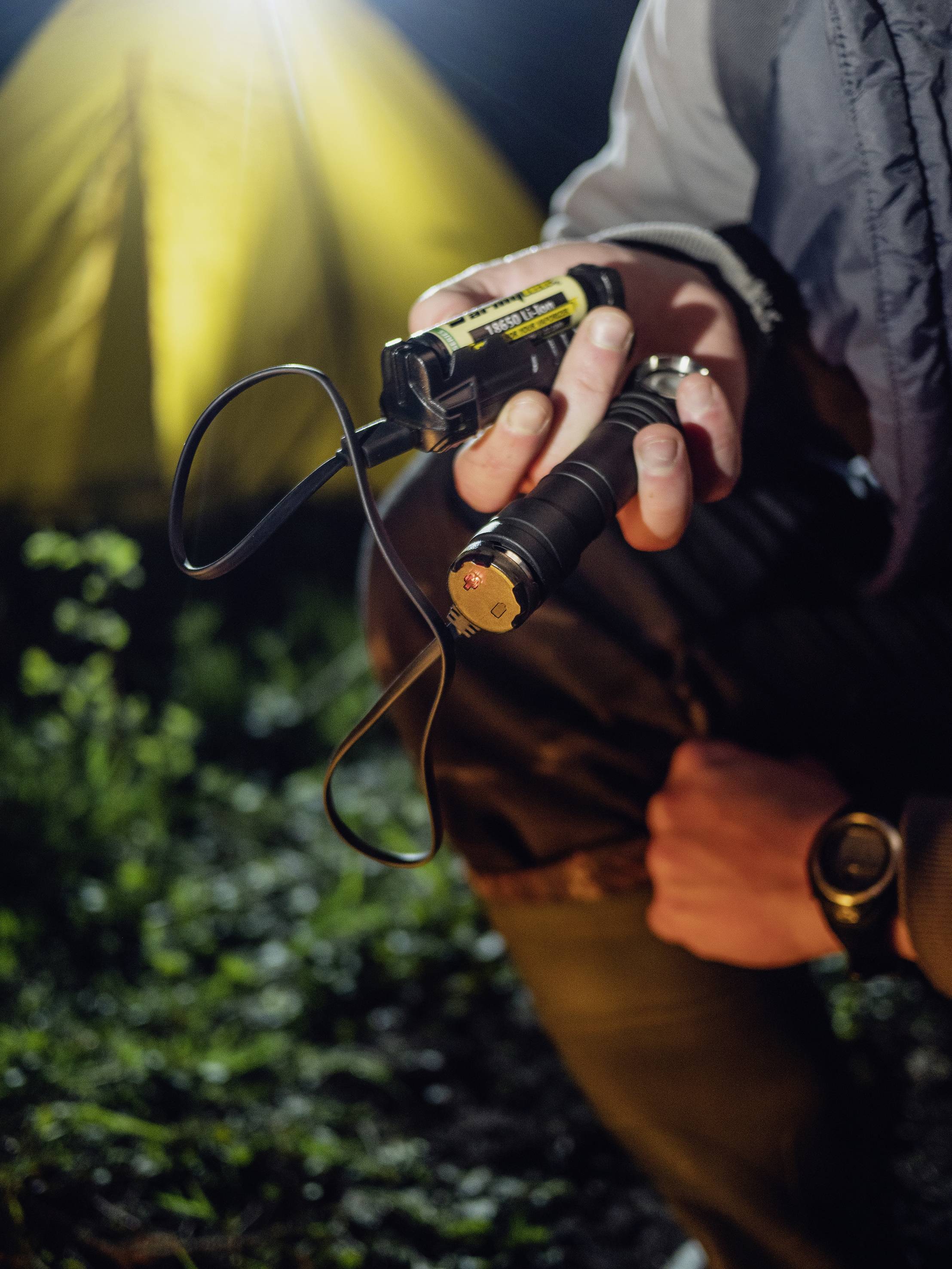 Eine Person hält eine Taschenlampe in der Hand. Im Hintergrund ist ein beleuchtetes Zelt bei Nacht zu sehen.