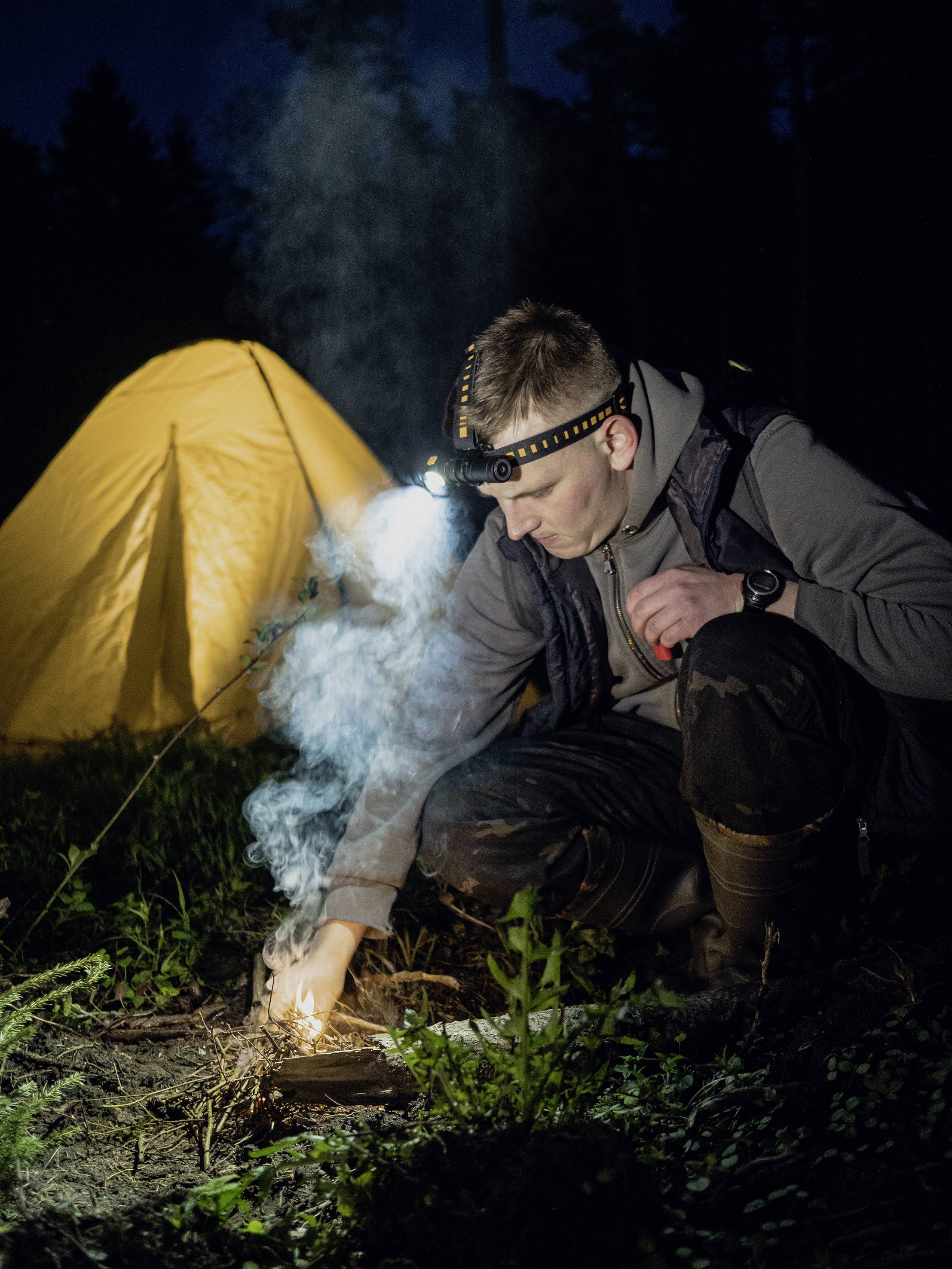 Eine Person mit Stirnlampe zündet abends ein Feuer an, im Hintergrund steht ein beleuchtetes gelbes Zelt.