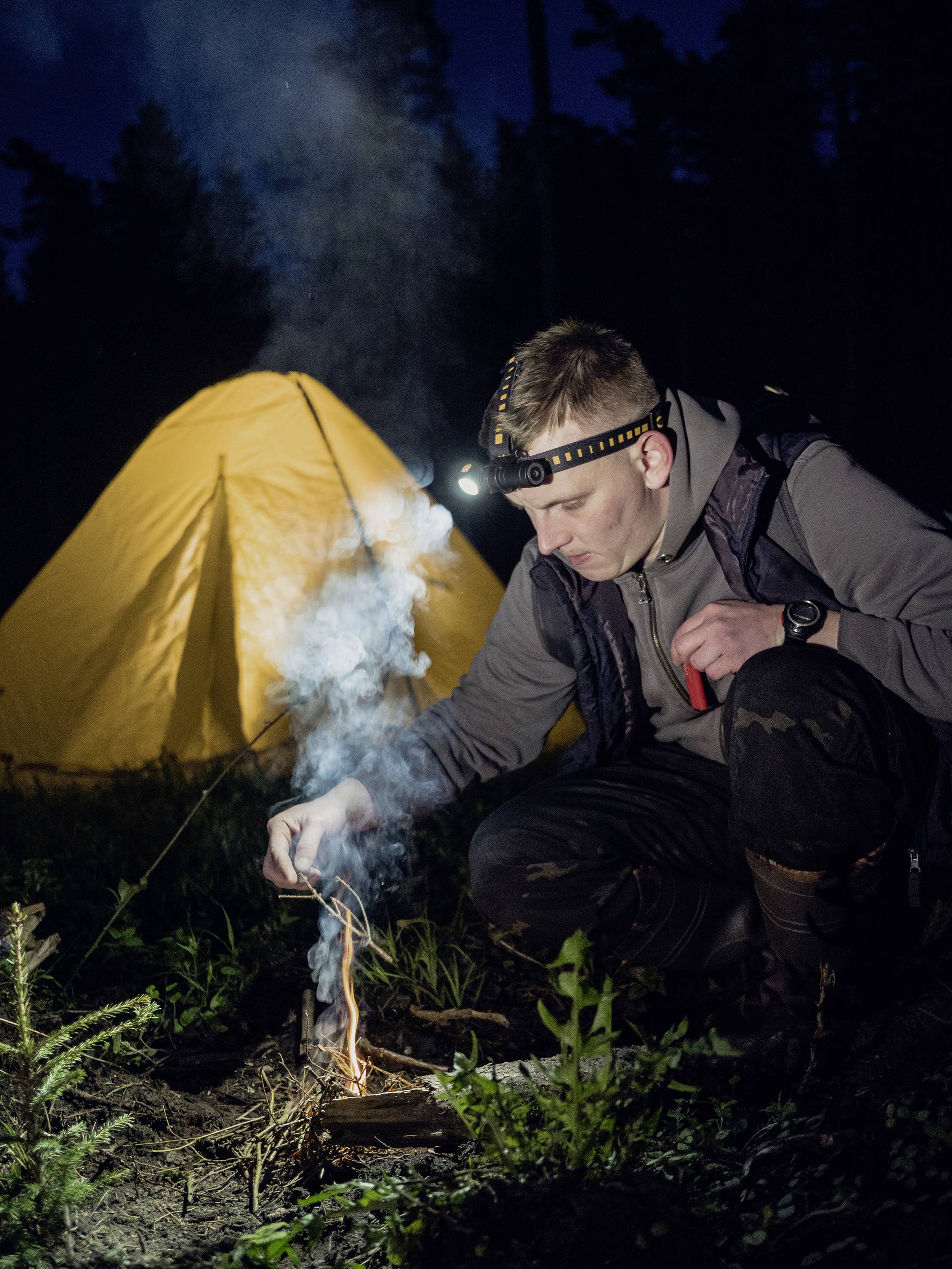 Ein Mann mit Stirnlampe zündet ein kleines Lagerfeuer an. Im Hintergrund steht ein beleuchtetes gelbes Zelt bei Nacht im Wald.