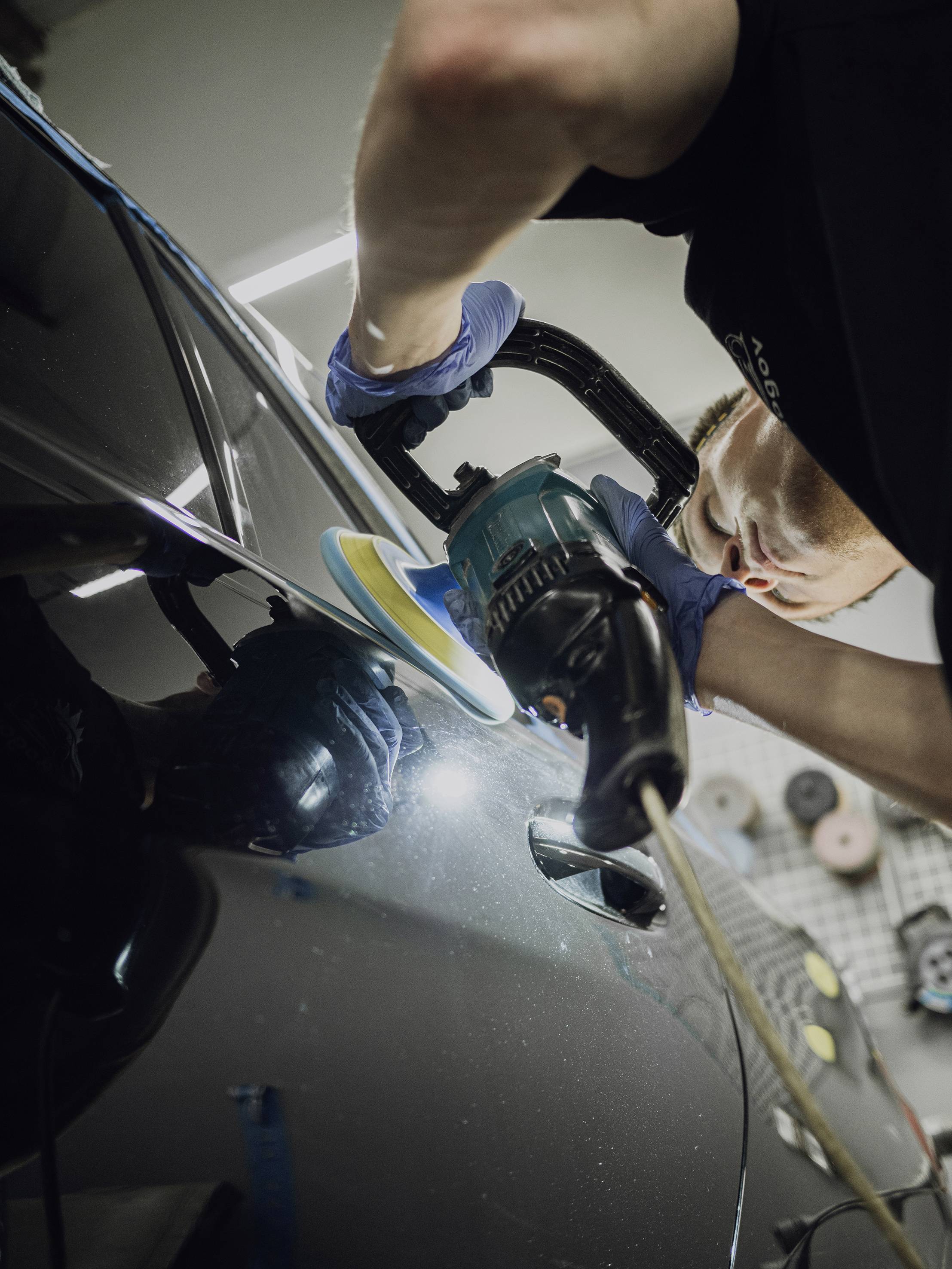 Eine Person poliert einen dunklen Autolack mit einer Maschine in einer Werkstatt, trägt dabei blaue Handschuhe.