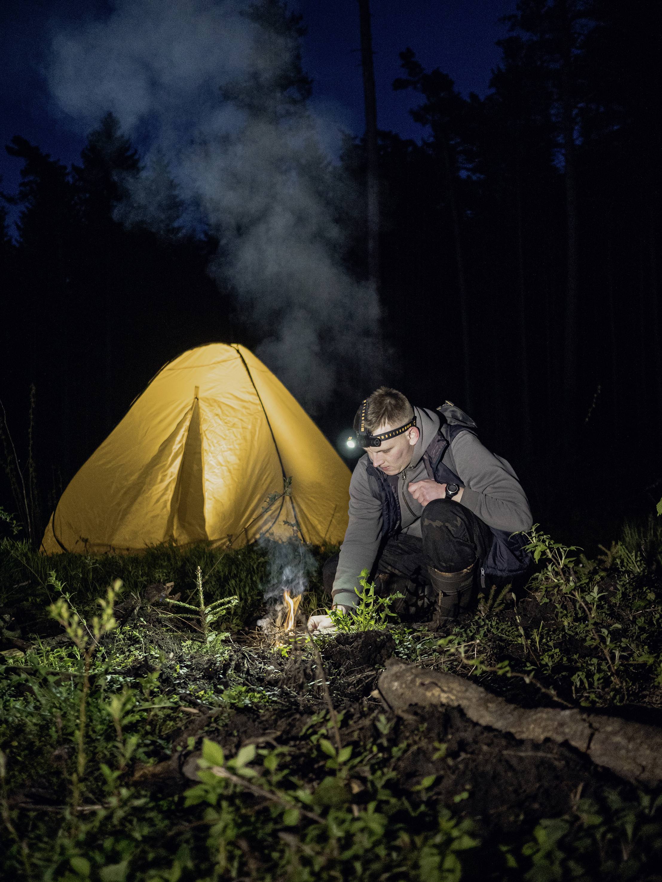Ein Mann entzündet ein Lagerfeuer vor einem gelben Zelt im dunklen Wald, trägt eine Stirnlampe; Nachtaufnahme.