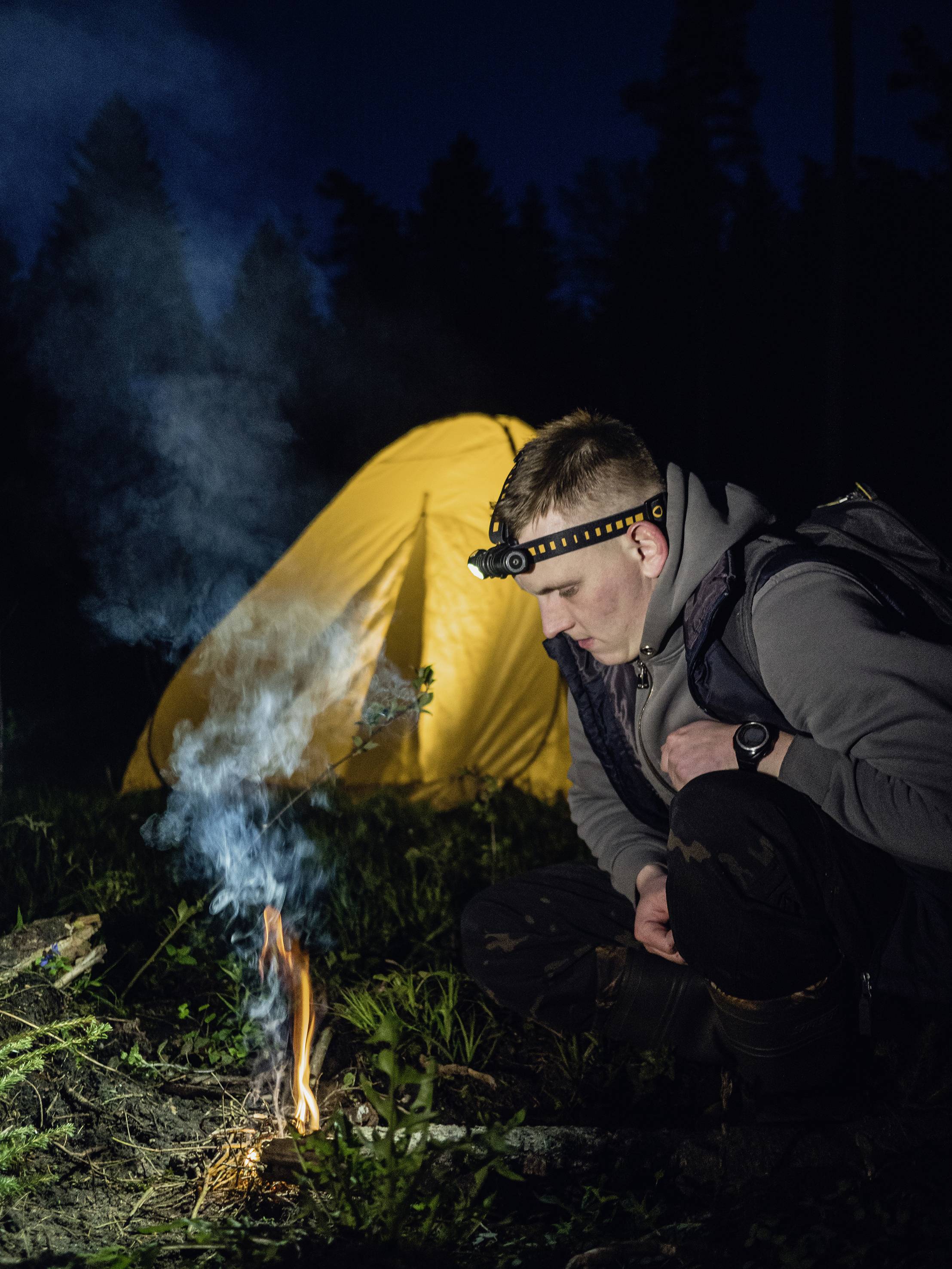 Ein Mann mit Stirnlampe hockt nachts neben einem kleinen Lagerfeuer, im Hintergrund ein beleuchtetes gelbes Zelt.
