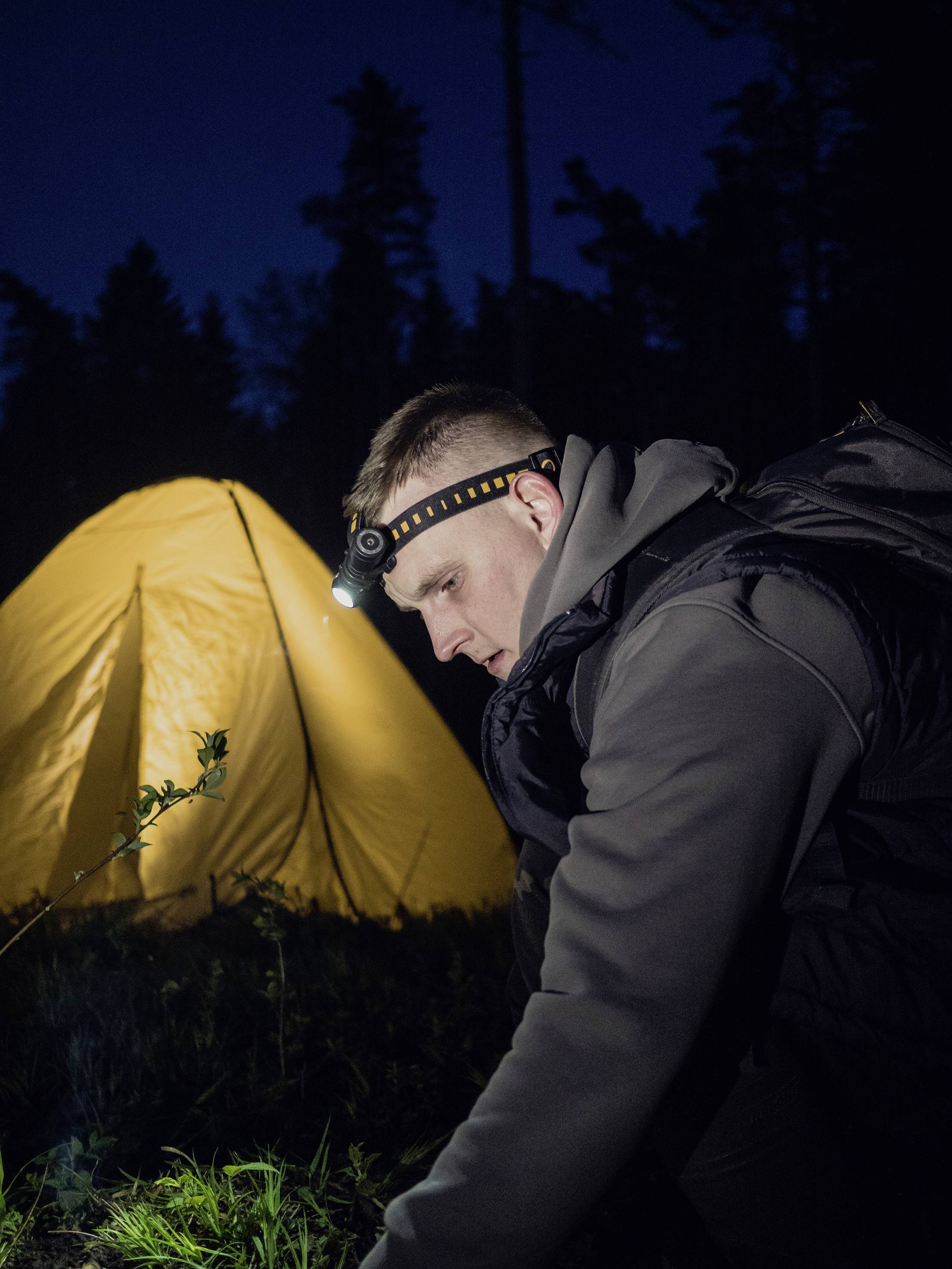 Ein Mann mit Stirnlampe kniet im Dunkeln vor einem leuchtend gelben Zelt im Wald und schaut konzentriert auf den Boden.