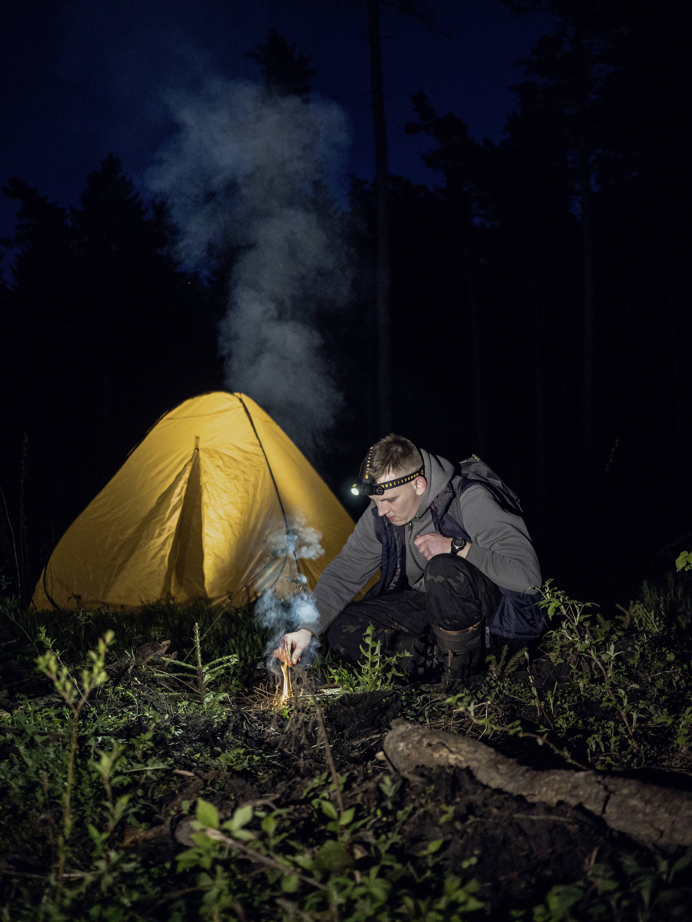 Eine Person entfacht ein kleines Feuer vor einem gelben Zelt in einem nächtlichen Wald. Sie trägt eine Kopflampe und ist in Outdoor-Kleidung.