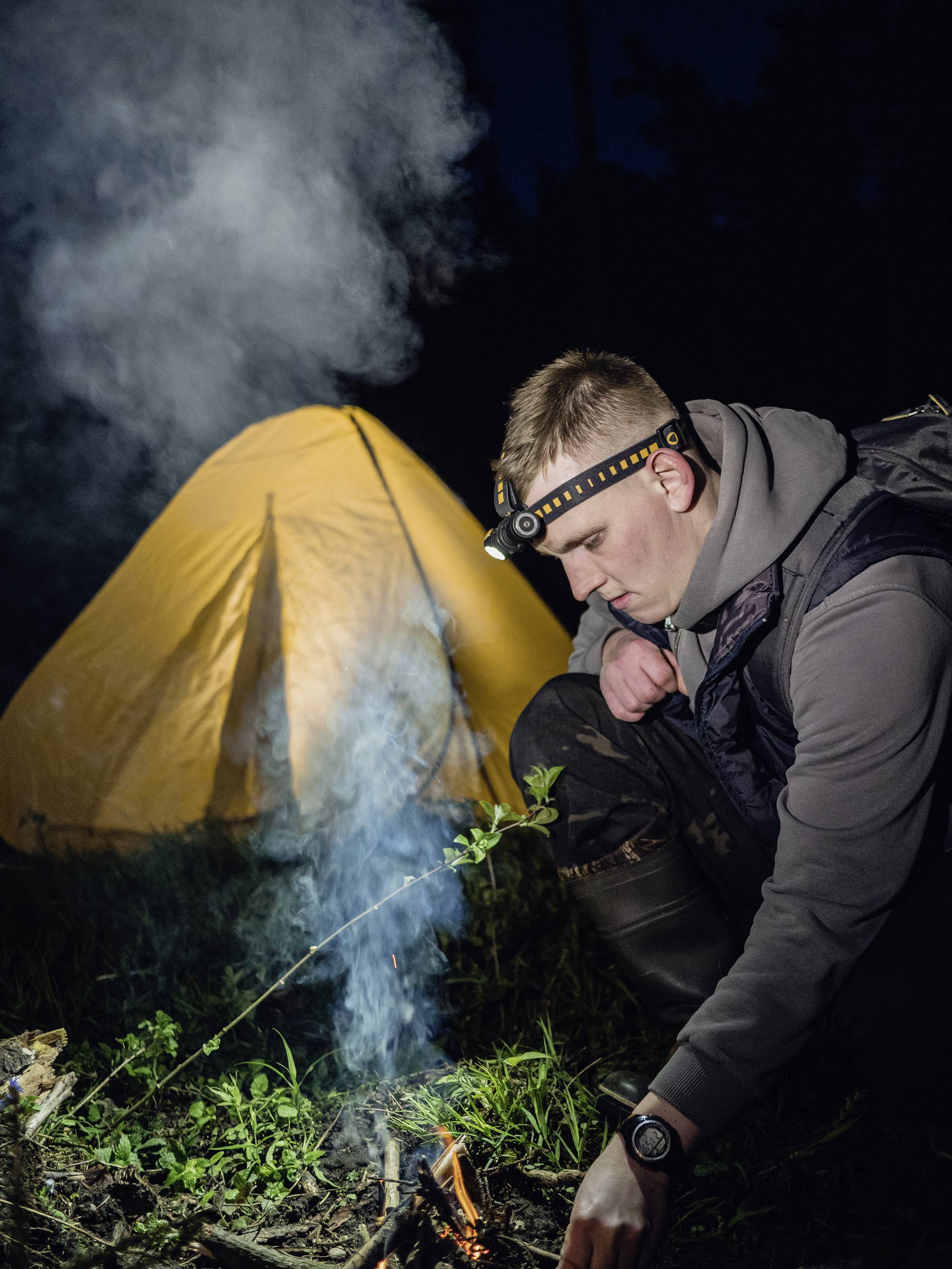 Ein Mann mit Stirnlampe kniet und entzündet ein Lagerfeuer. Im Hintergrund steht ein beleuchtetes gelbes Zelt im Dunkeln.