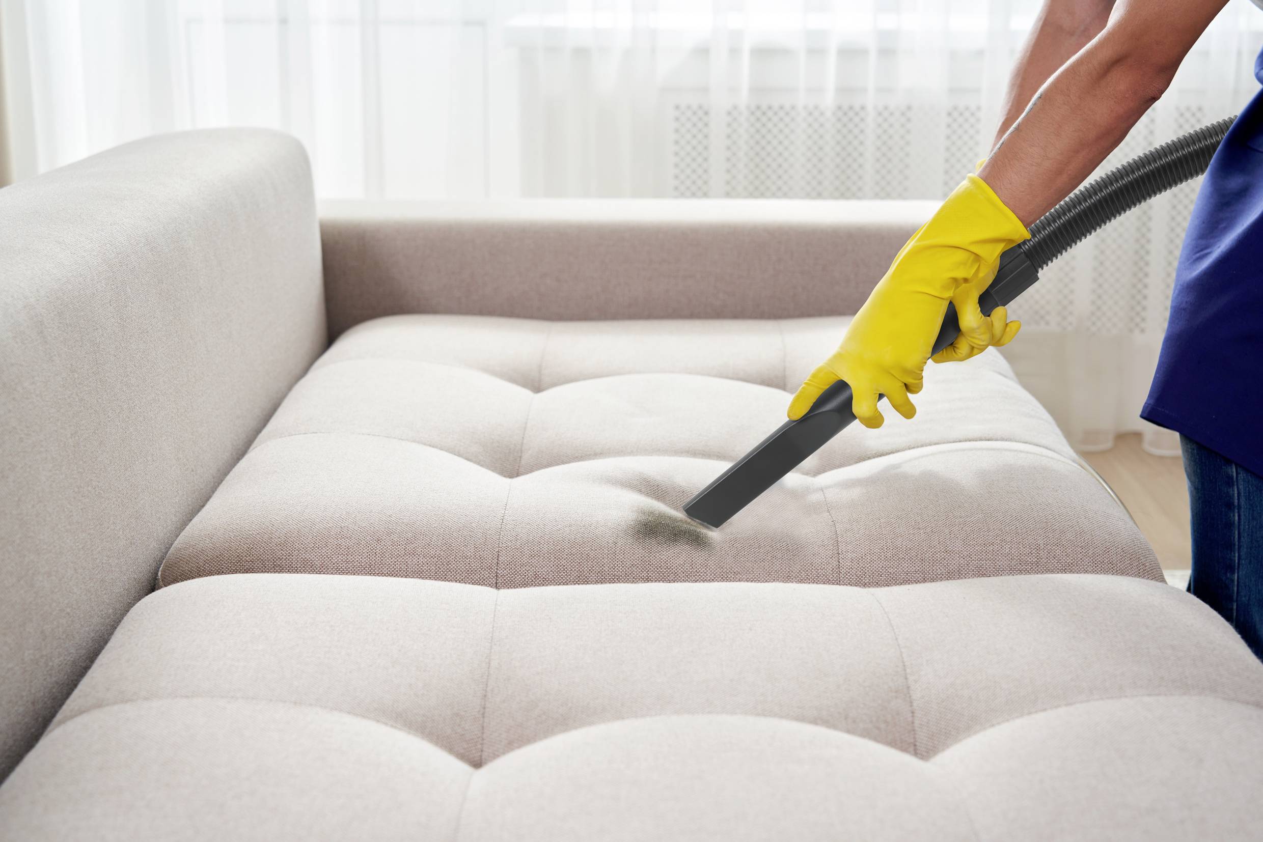 Eine Person reinigt ein beigefarbenes Sofa mit einem Staubsauger. Sie trägt gelbe Reinigungshandschuhe.