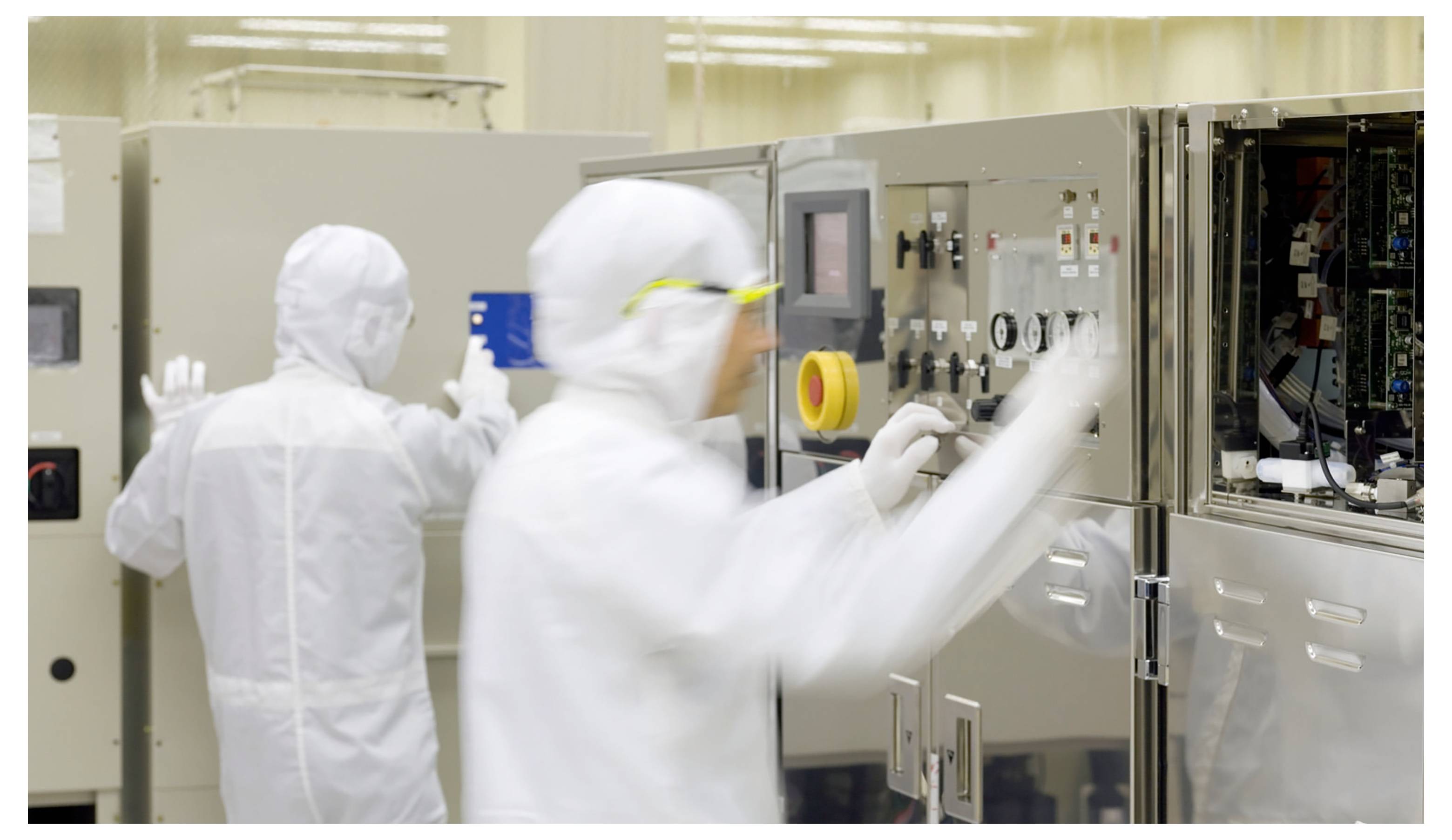 Zwei Techniker in Reinraumumzügen bedienen Halbleiterausrüstung in einem Labor, wobei einer die Steuerungen einstellt und der andere an einer nahegelegenen Station arbeitet.