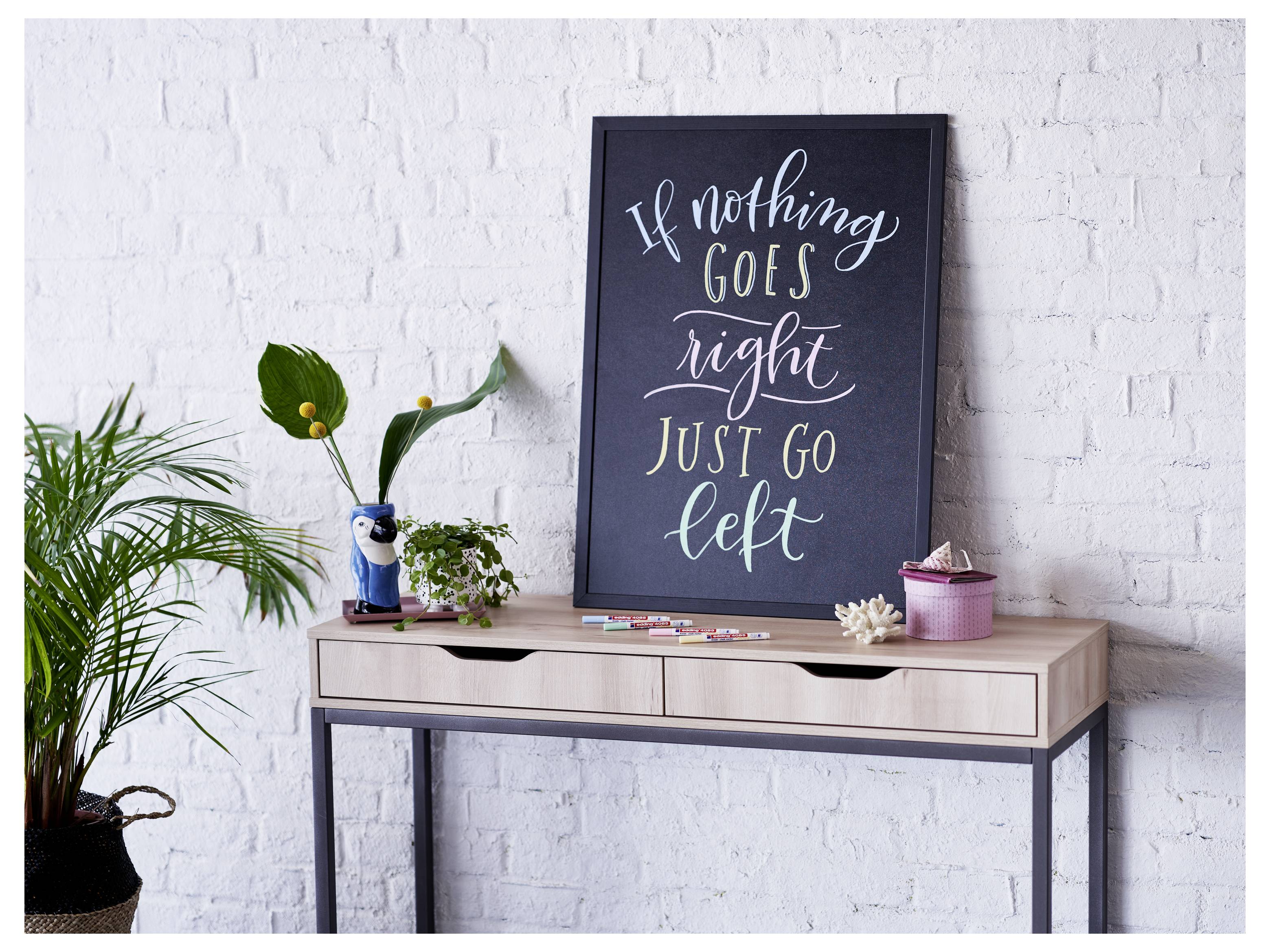 Ein gerahmtes Motivationsschild auf einem Tisch mit der Aufschrift „Wenn nichts nach rechts geht, geh einfach nach links", umgeben von Pflanzen und kleiner Dekoration gegen eine weiße Backsteinwand.