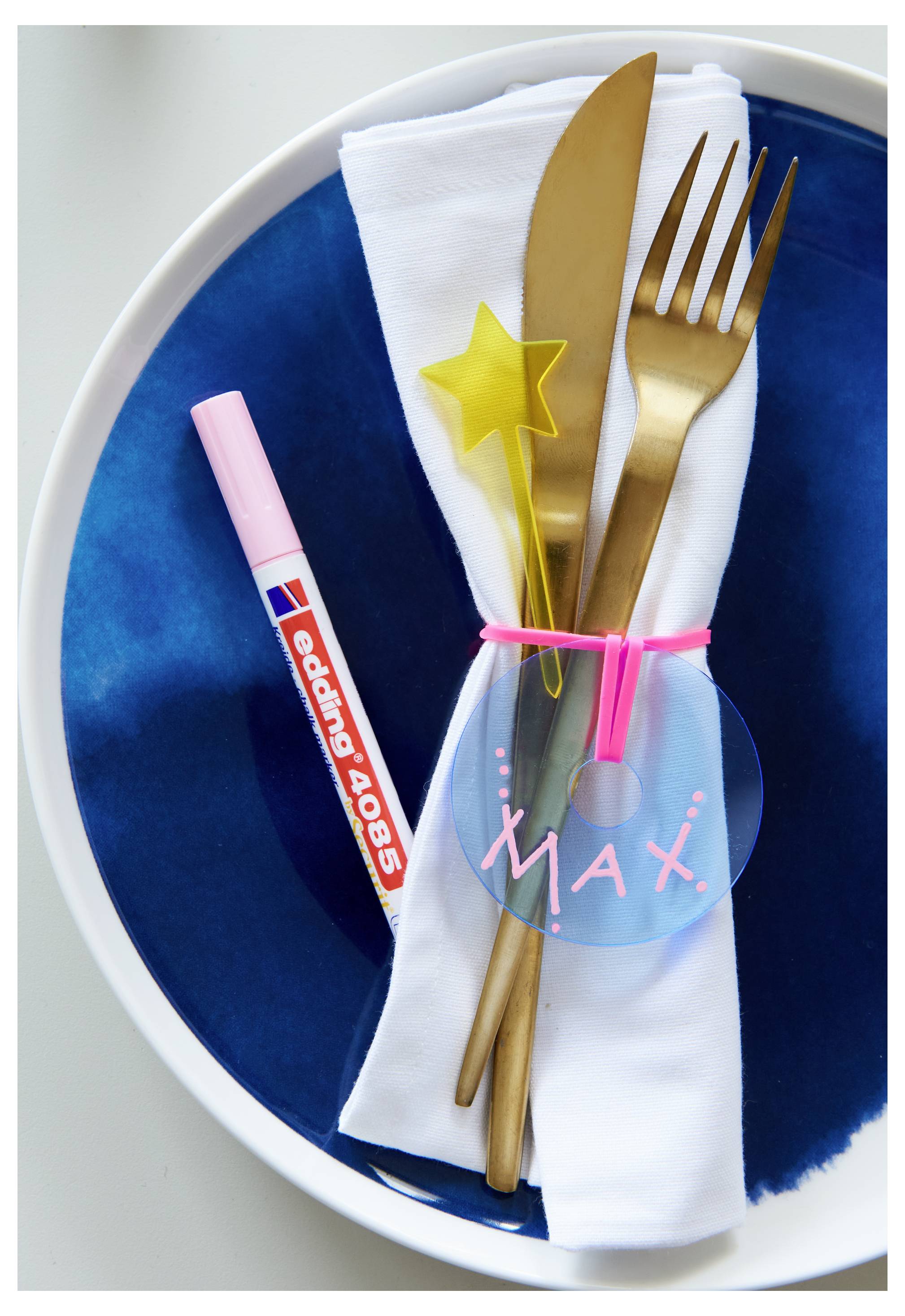 Gedeck mit gefaltetem weißem Serviette auf blauem Teller, gebunden mit einem „Max"-Namensschild und rosa Schleife, plus goldenen Bestecken und einem Stern-Rührer; ein pink-gestreifter Stift liegt daneben.