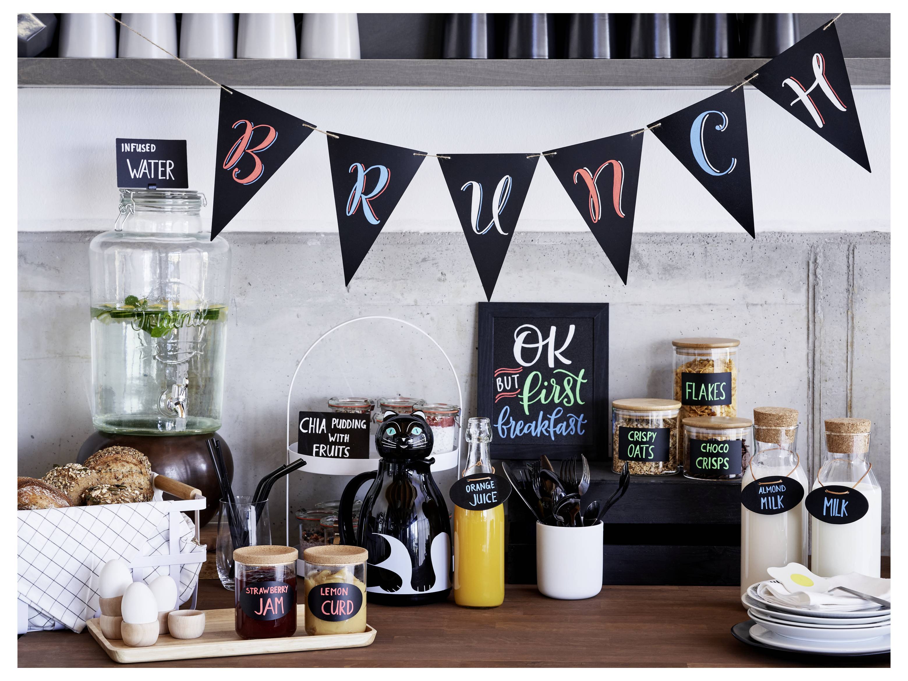 Frühstücksbuffet-Display mit „BRUNCH"-Banner, Wasserspender, Gebäck, Gläser mit Würzmitteln, Getreide, Saft, Milch und Tellern auf einer Theke.