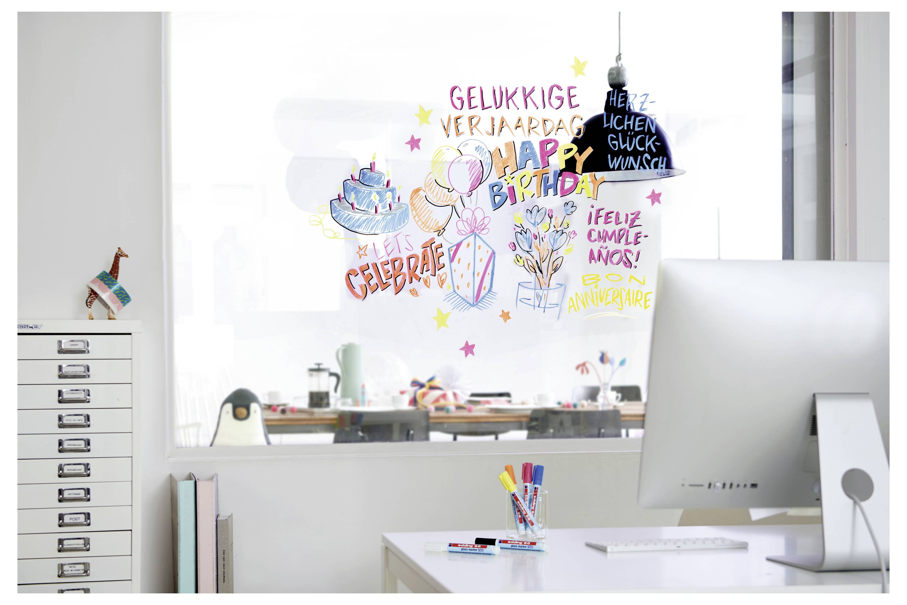 Schreibtisch im Büro mit Whiteboard, das mit bunten „Alles Gute zum Geburtstag"-Grüßen in mehreren Sprachen und Geburtstagszeichnungen bedeckt ist.