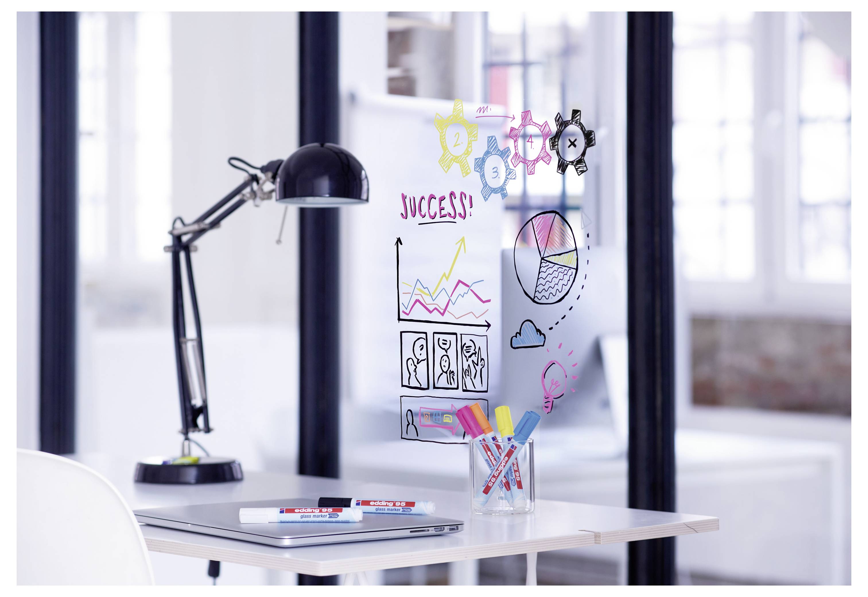 Büroschreibtisch mit Laptop, Lampe und einer Glastafele, auf der "ERFOLG!" mit Diagrammen, Symbolen und Skizzen zum Brainstorming und zur Planung zu sehen ist.