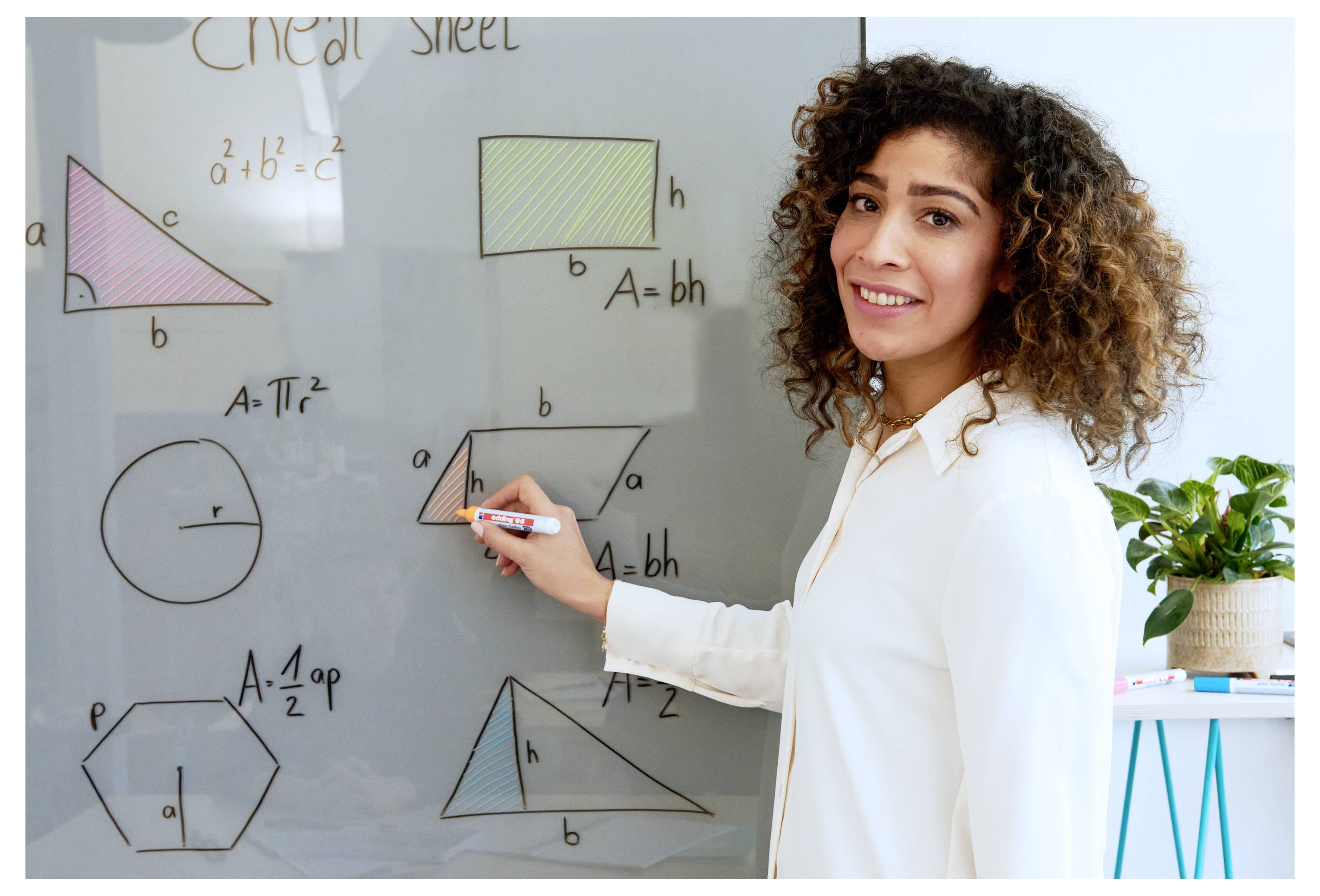 Lächelnde Lehrerin zeigt auf ein Geometrie-Whiteboard mit Flächenformeln und Formen und unterrichtet Mathematik in einem Klassenzimmer.