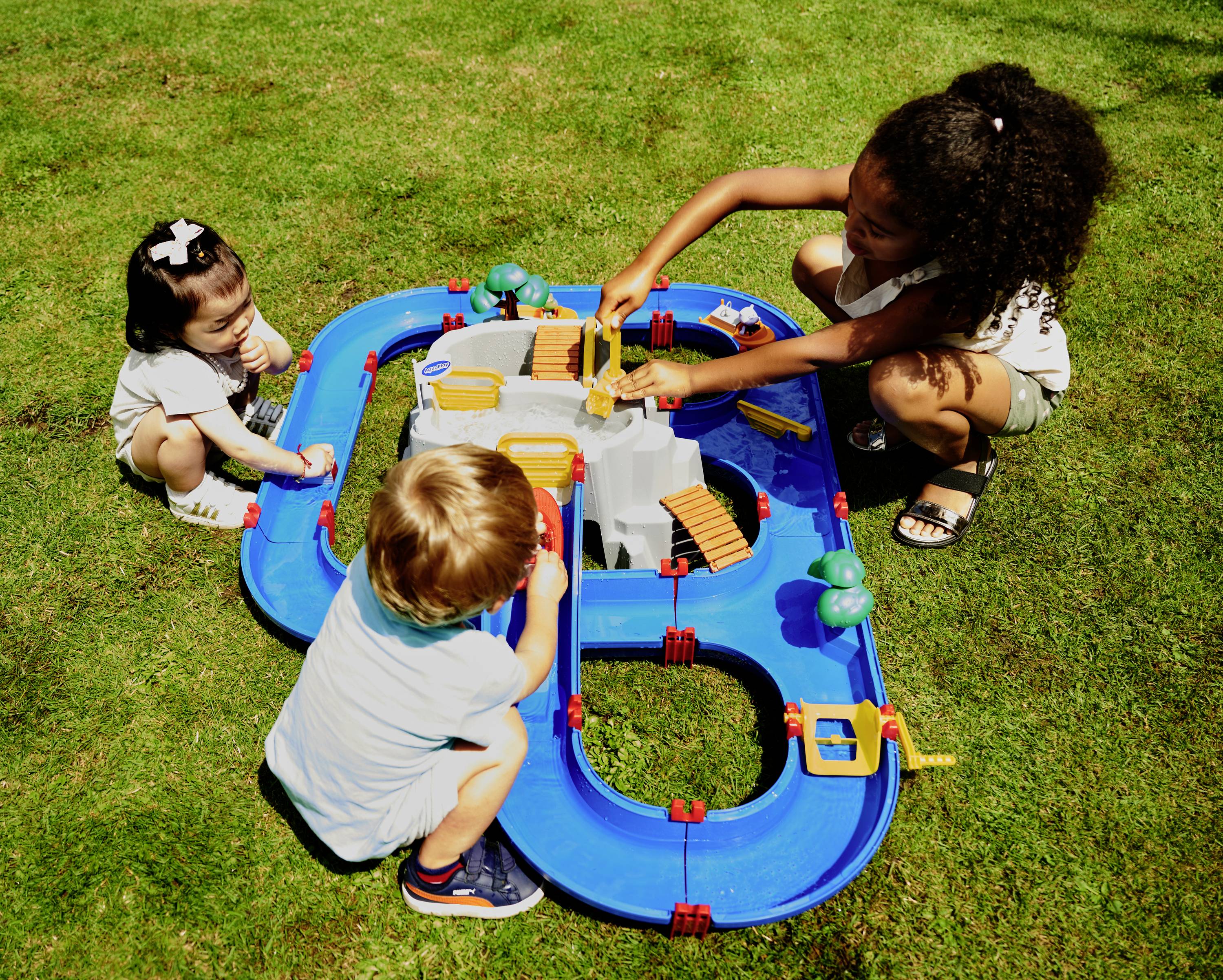 Drei Kinder spielen draußen auf einer Wiese mit einem blauen Wasserspielzeug, das Kanäle und eine kleine Burg enthält.