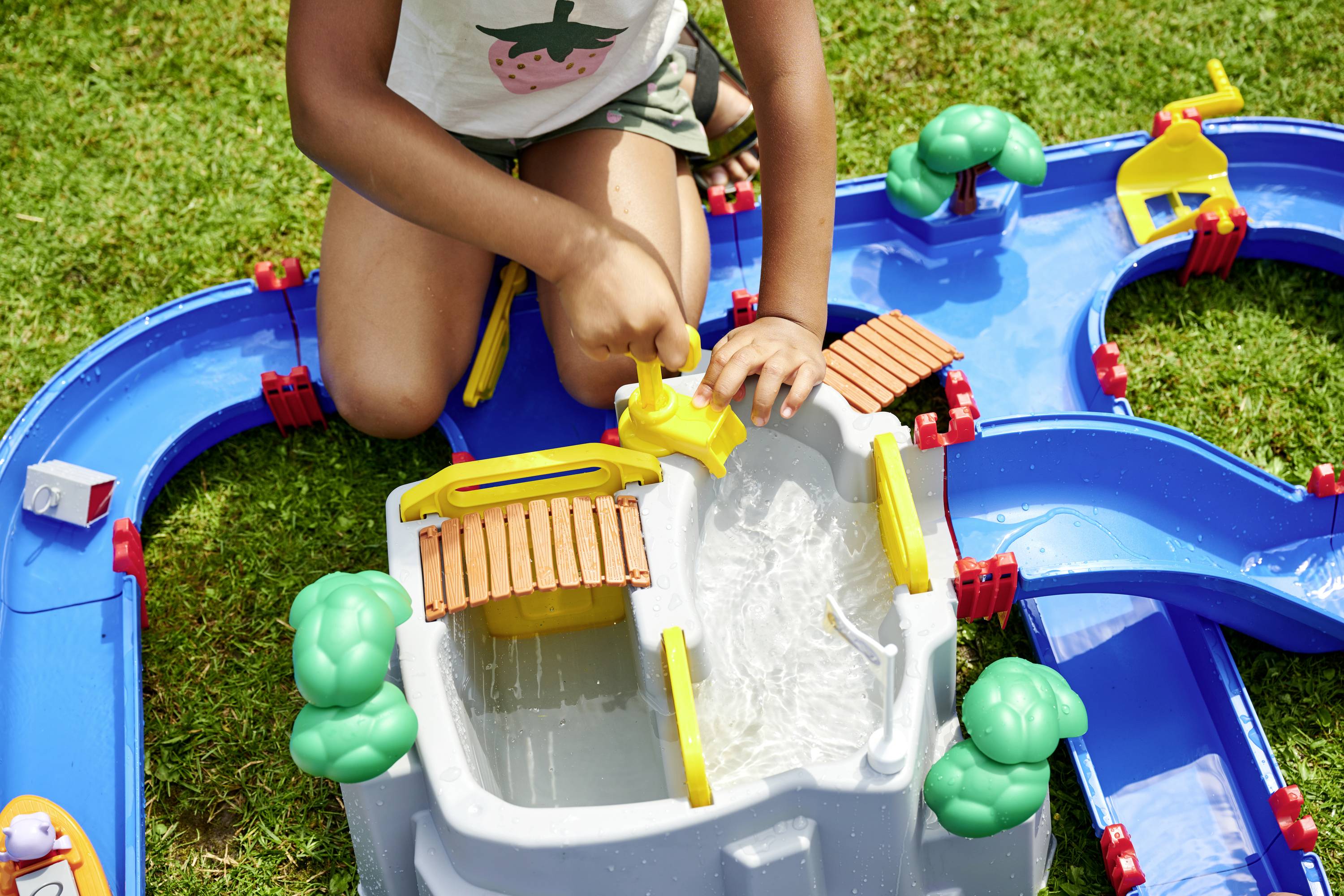 Ein Kind spielt mit einem blauen Wasserspielzeug, das über Wasserwege und Schleusensysteme verfügt, im Freien auf Rasen.