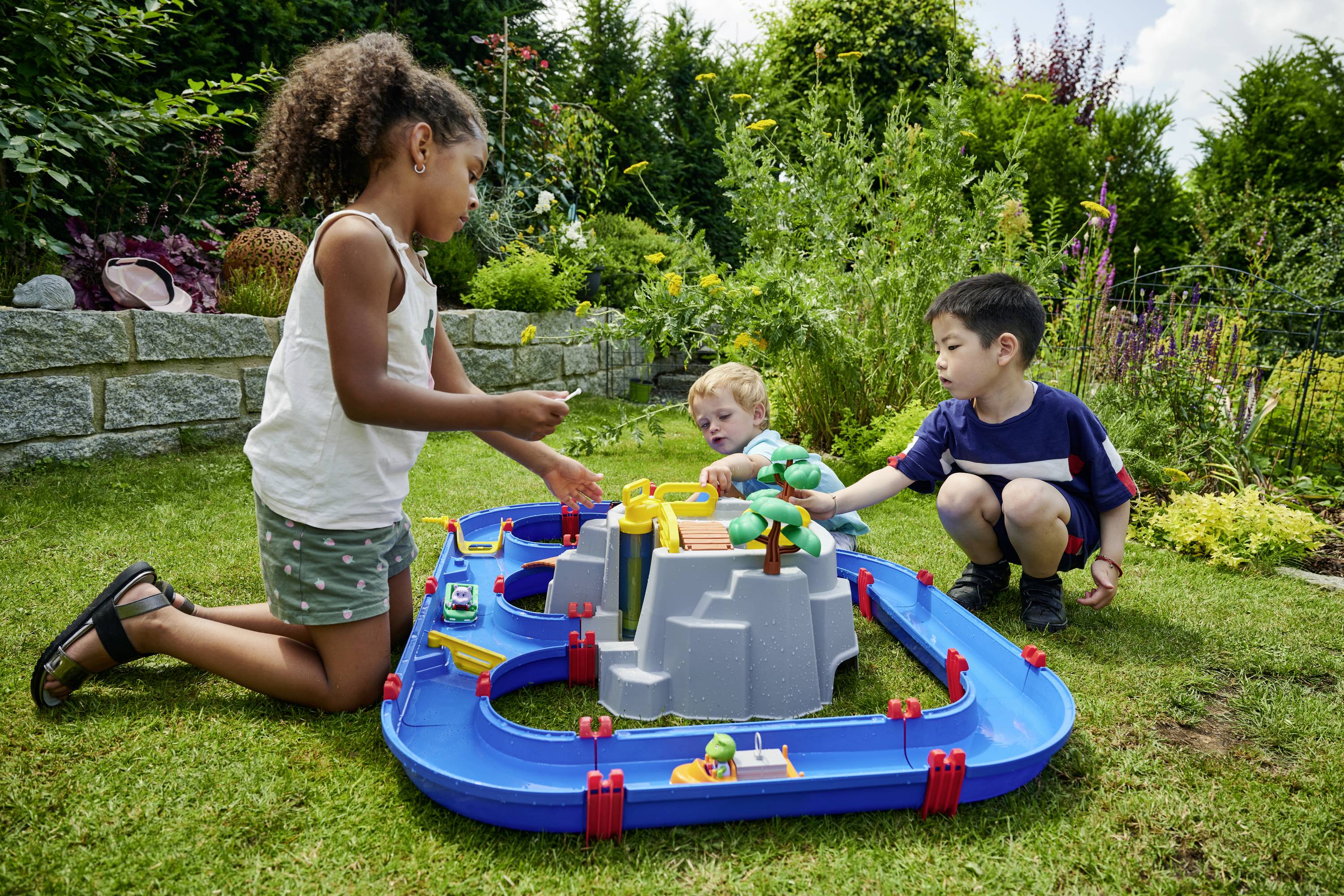 Drei Kinder spielen im Garten mit einer Spielzeugbahn, die um eine graue zentrale Struktur verläuft. Sie sind in grüner Umgebung.