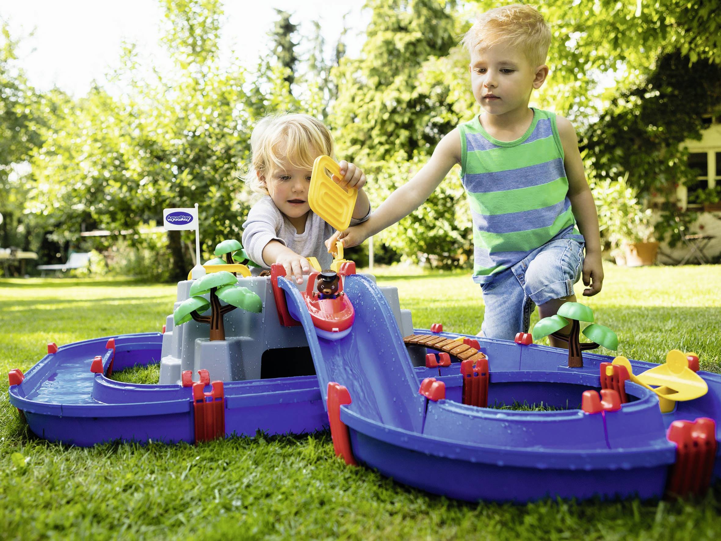 Zwei Kinder spielen draußen mit einem blauen Wasserbahn-Set. Sie gießen Wasser hinein, während Plastikbäume um den Kanal stehen.