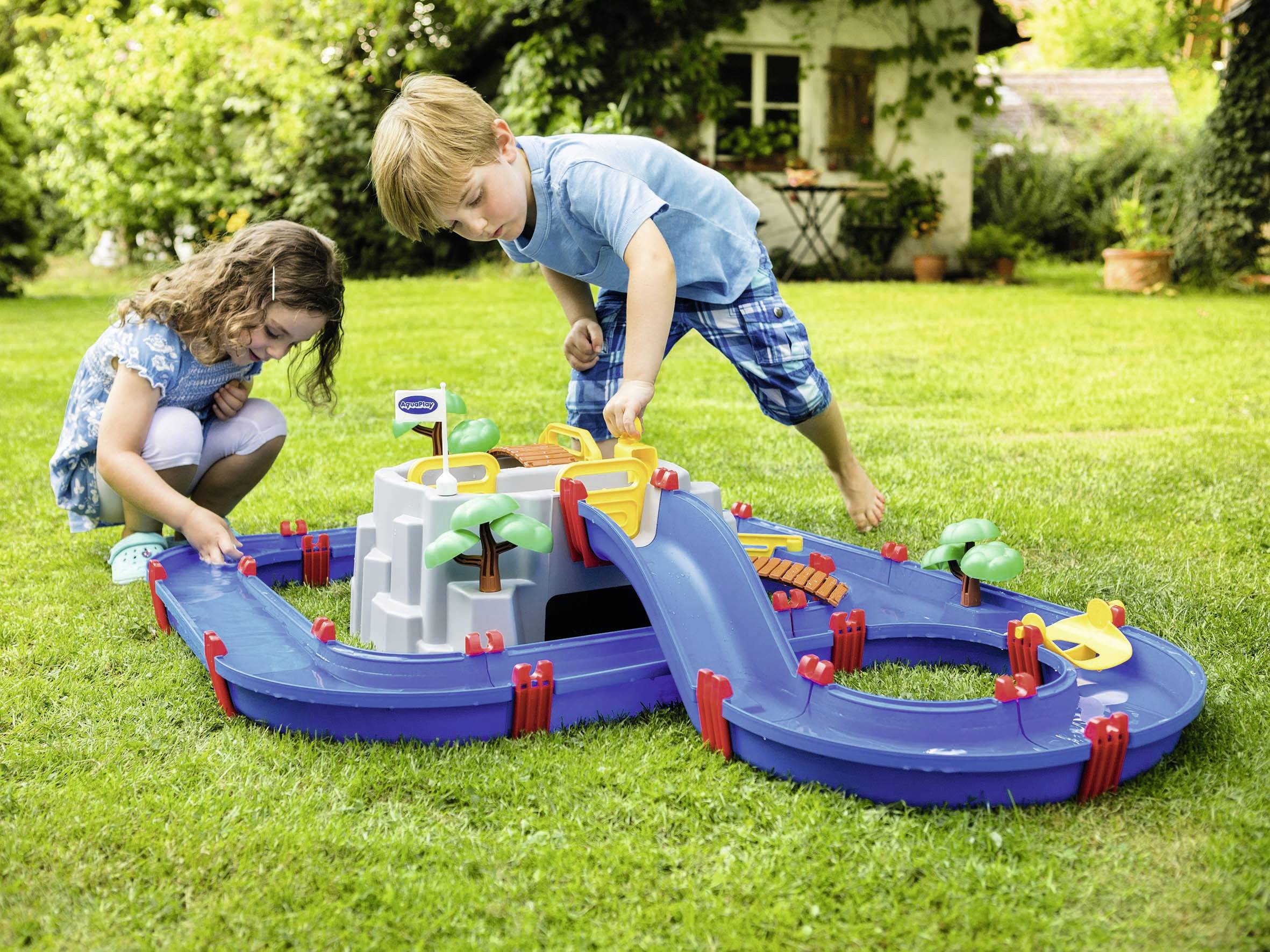 Zwei Kinder spielen im Garten mit einer Wasserbahn. Das Set enthält Kanäle, Brücken und kleine Boote, die im Wasser schwimmen.