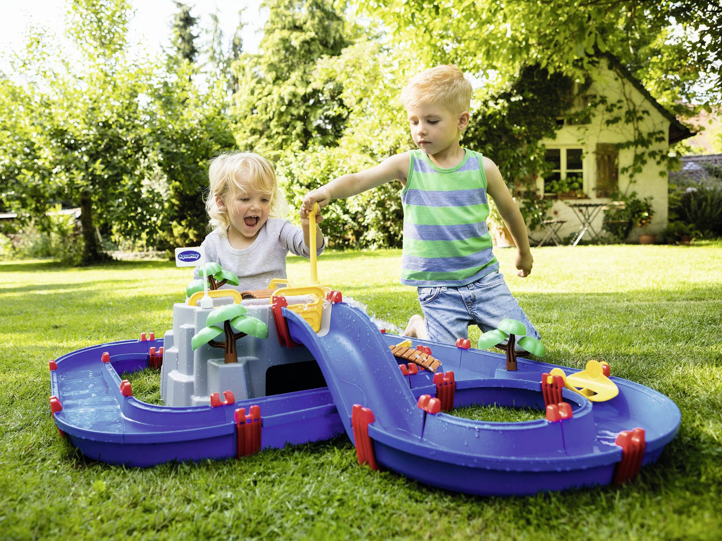 Zwei Kinder spielen im Garten mit einer blauen Wasserspielbahn. Ein Junge trägt gestreiftes Shirt, das andere Kind freut sich am Spiel. Im Hintergrund steht ein Haus.