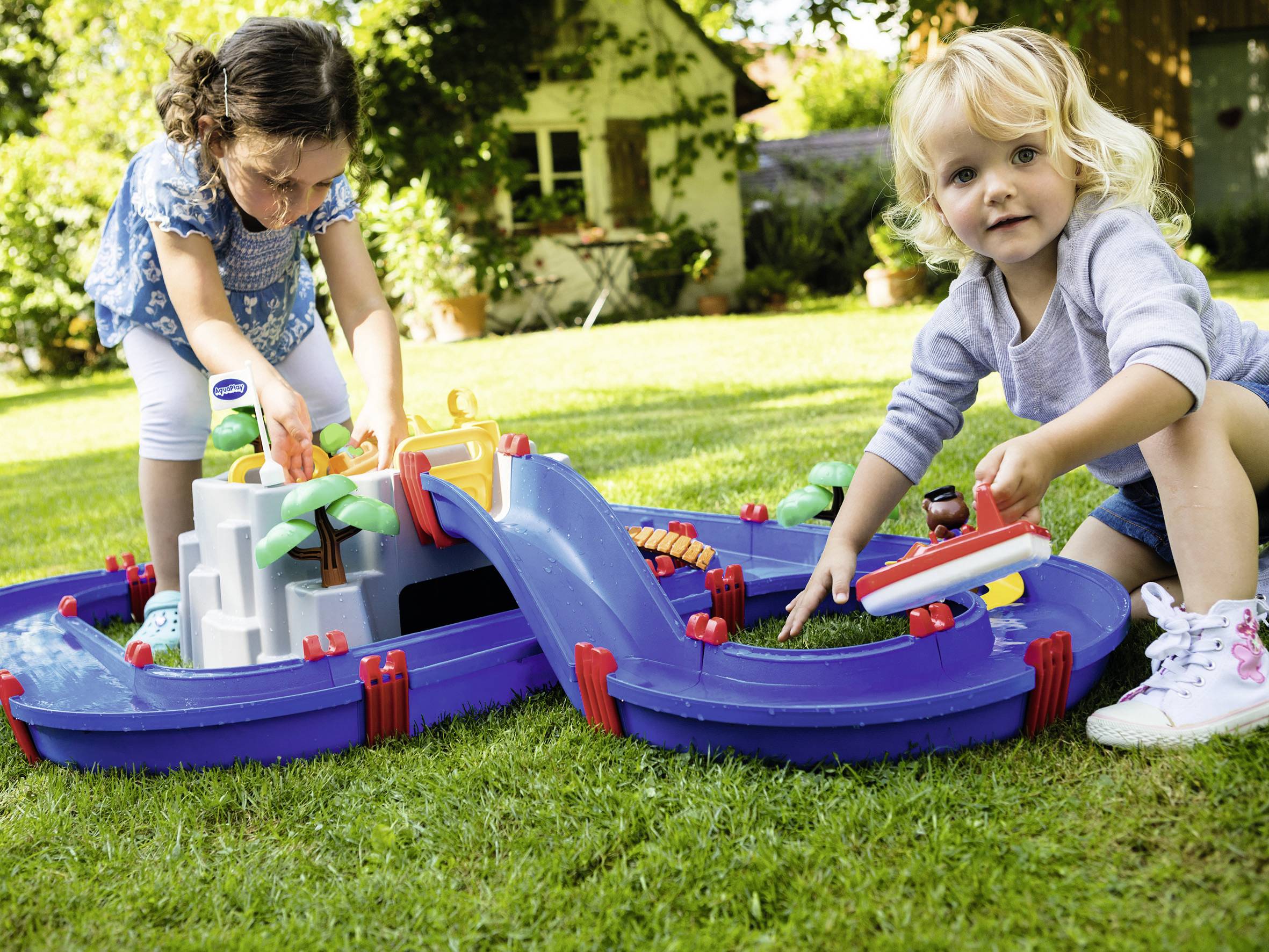 Zwei Kinder spielen im Garten mit einer blauen Wasserbahn, die Palmen und Boote enthält. Im Hintergrund ist ein Haus zu sehen.