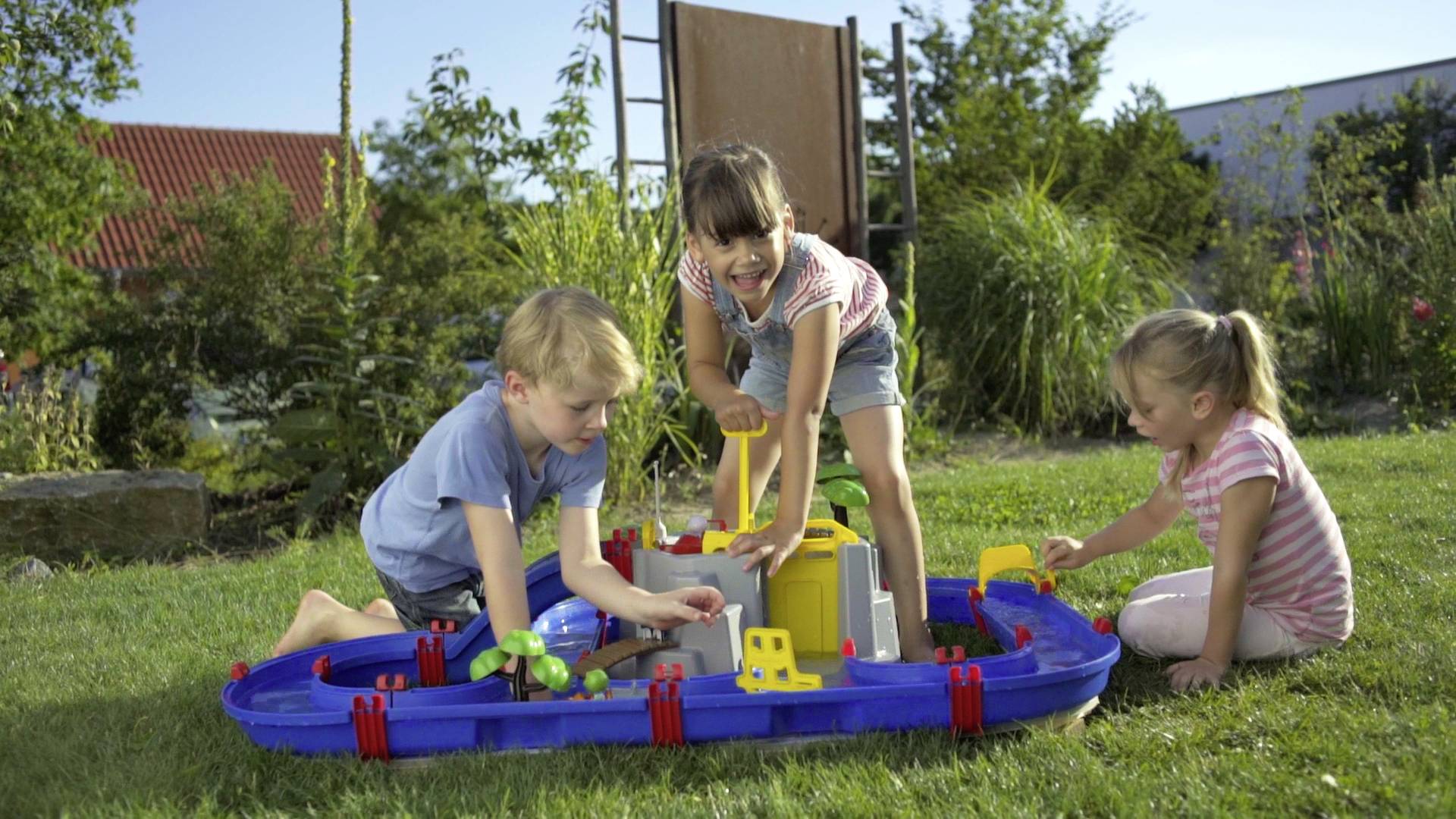 Drei Kinder spielen draußen auf einer Wiese mit einer blauen Wasserspielbahn. Im Hintergrund sind Bäume und Gebäude zu sehen.