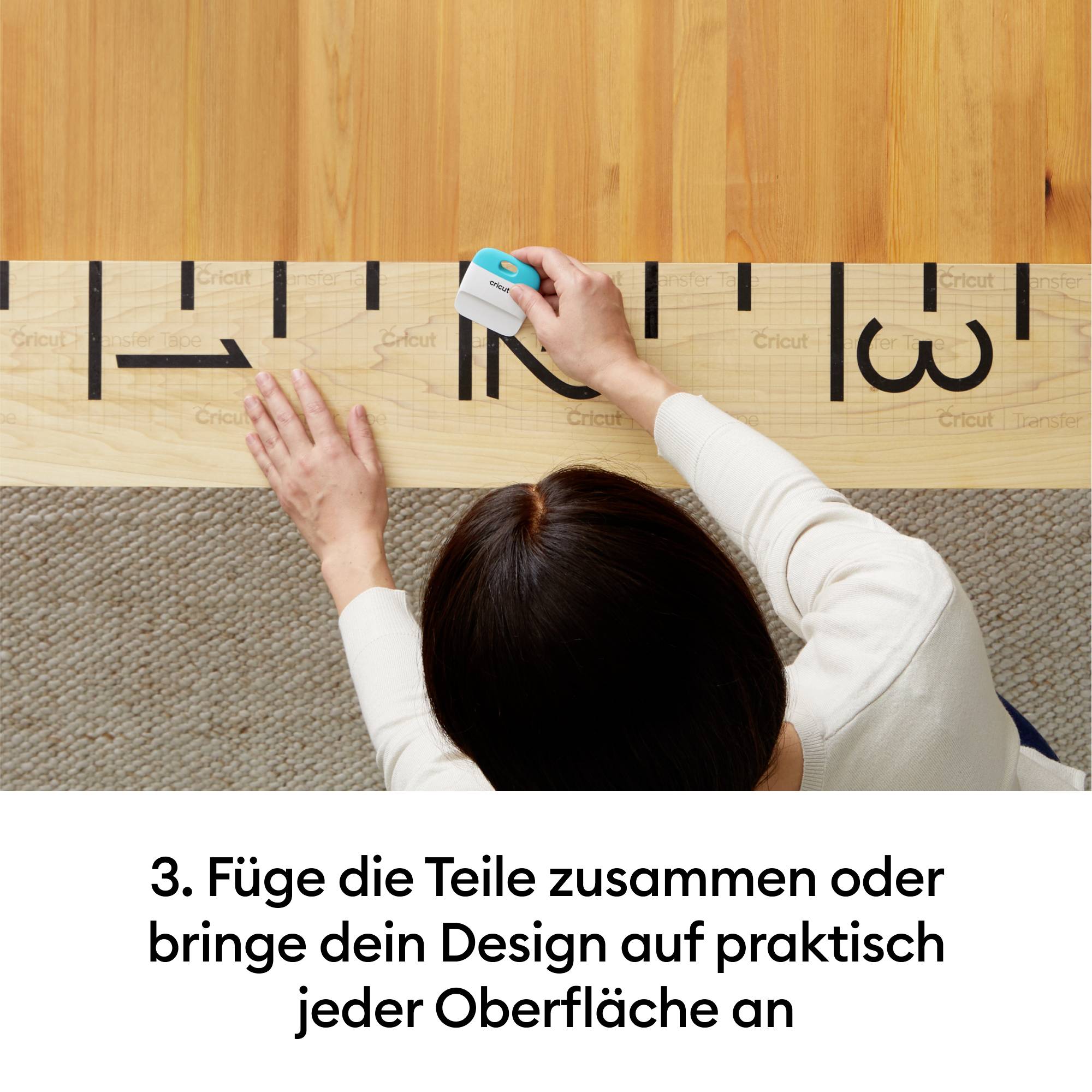 Person klebt ein Design mit einem Tool auf eine Holzoberfläche. Text darunter: '3. Füge die Teile zusammen oder bringe dein Design auf praktisch jeder Oberfläche an'.