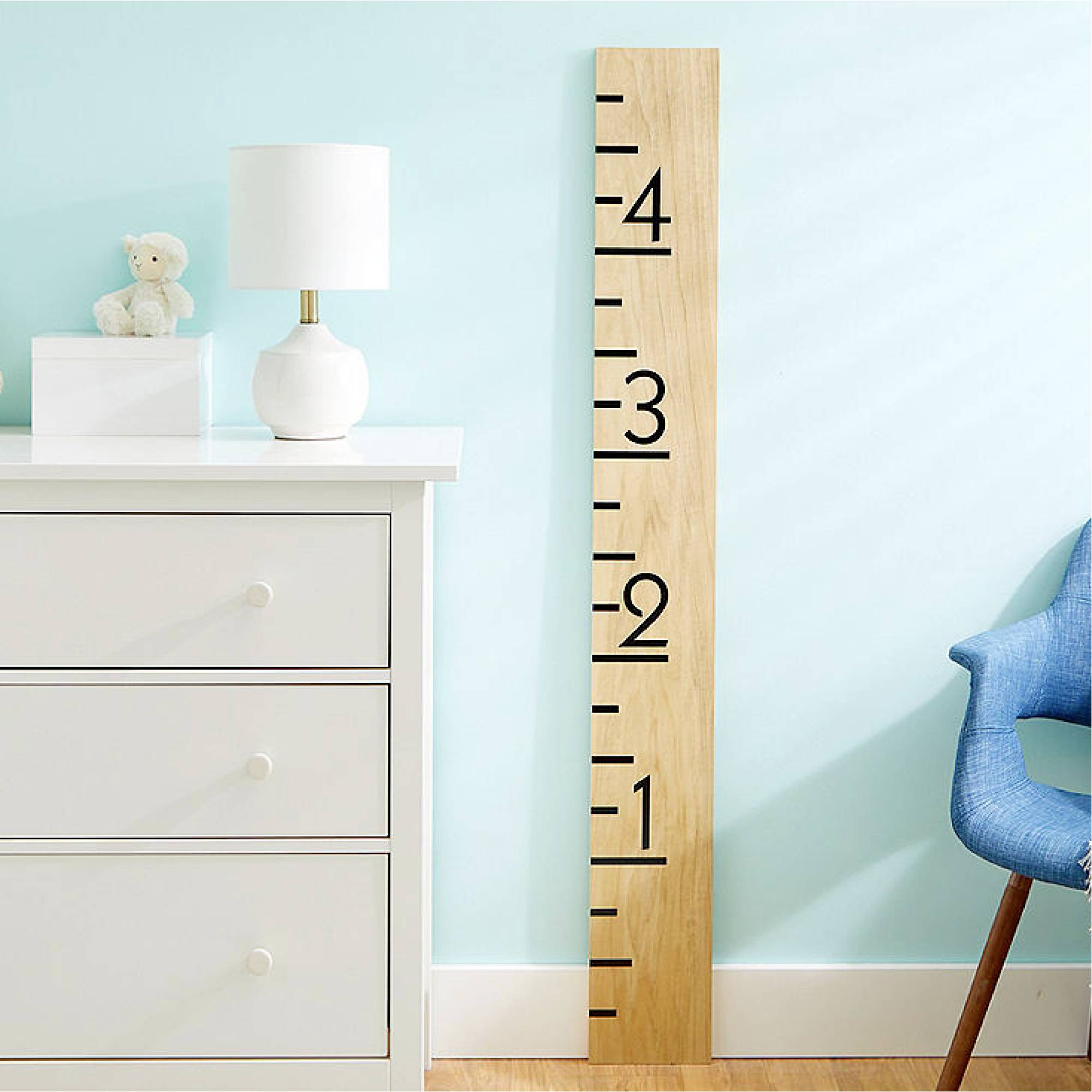 Helles Kinderzimmer mit weißer Kommode, blauer Wand und Holzwachstumsdiagramm daneben; ein Teddybär und Lampe auf der Kommode.