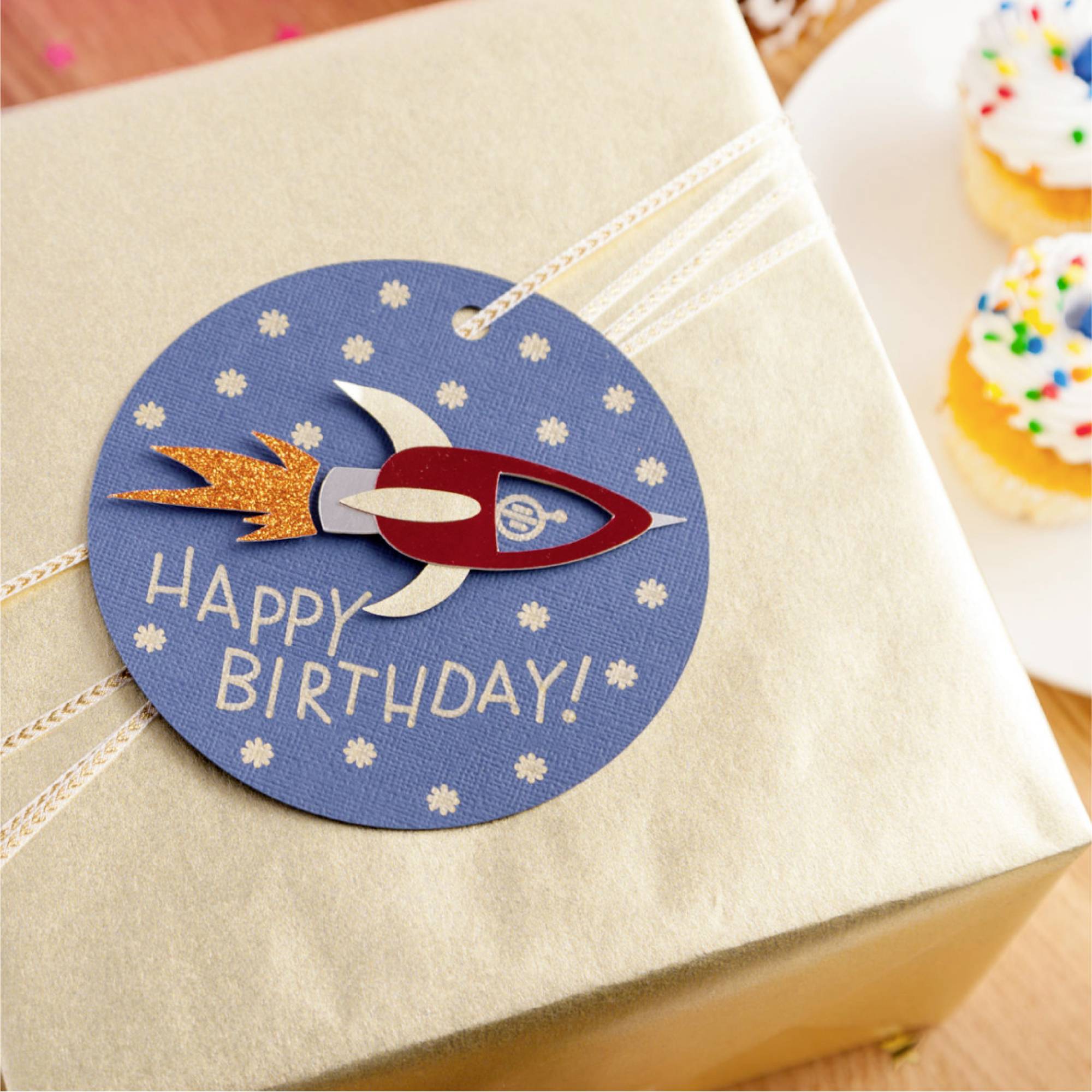 Geschenk mit blauer Geburtstagskarte, Raketenmotiv und 'Happy Birthday'-Schriftzug, auf Holztisch mit zwei Cupcakes im Hintergrund.