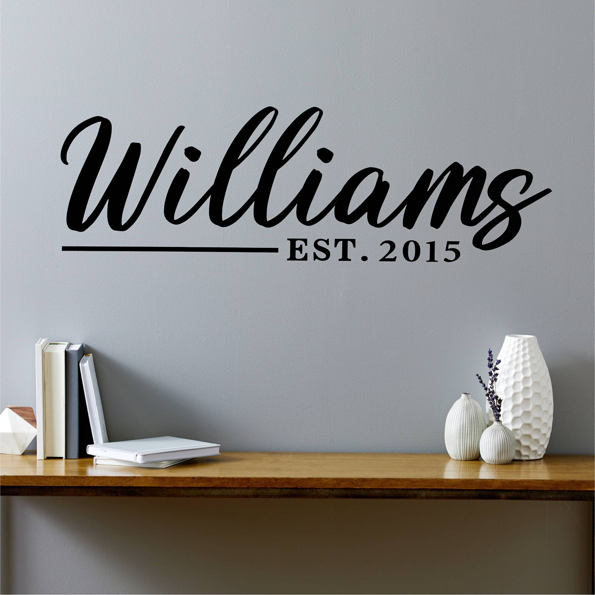 'Williams. EST. 2015.' Schriftzug an grauer Wand über Holzschreibtisch mit Dekorationen: Büchern, Vasen, Topfpflanze.