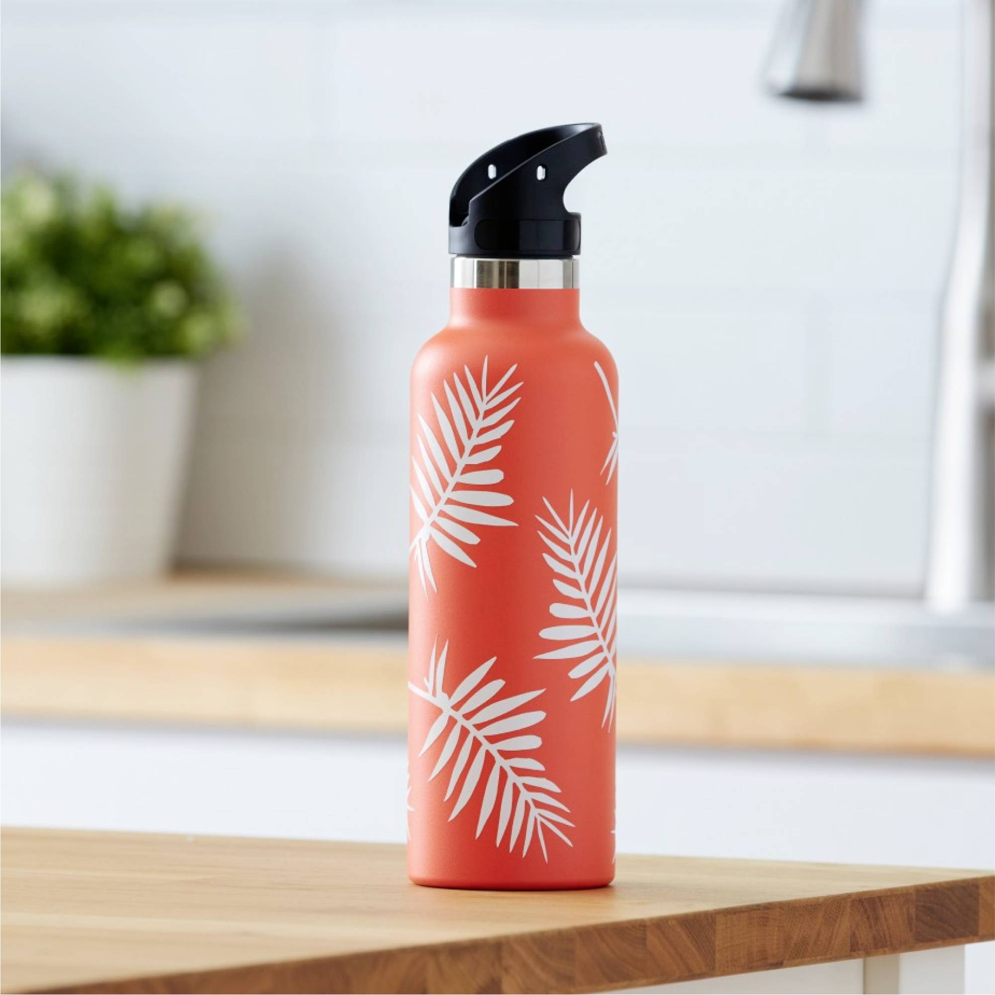 Eine orangefarbene Trinkflasche mit weißem Palmenblätter-Muster steht auf einer Holzoberfläche in einer Küche.