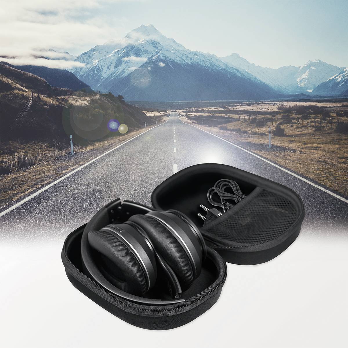 Ein Paar schwarze Kopfhörer liegt in einem geöffneten Etui auf einer Straße mit schneebedeckten Bergen im Hintergrund.