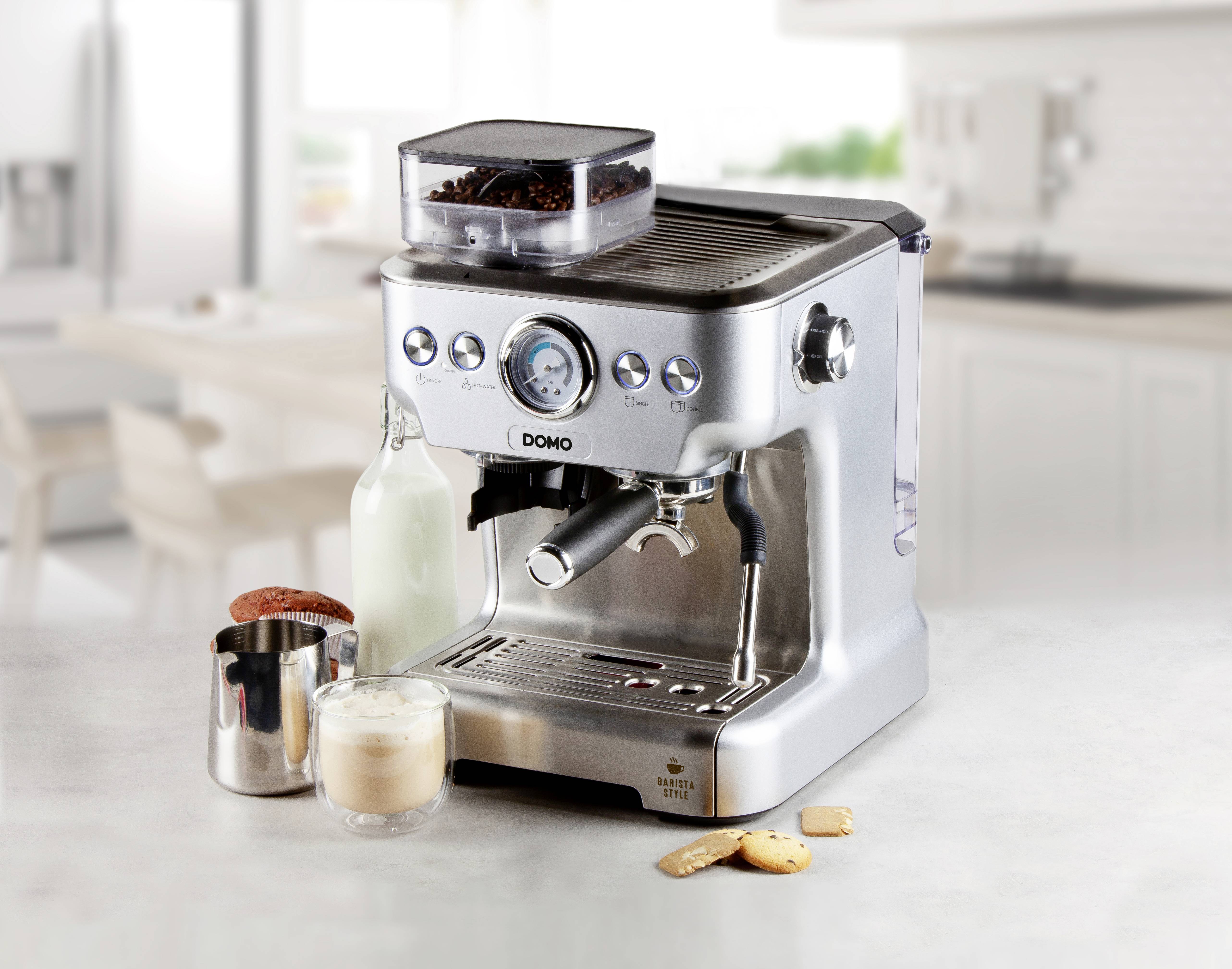 Eine silberne Espressomaschine auf einer Küchenarbeitsplatte, umgeben von einem Milchkännchen, einem Glas Milchschaum und Keksen.