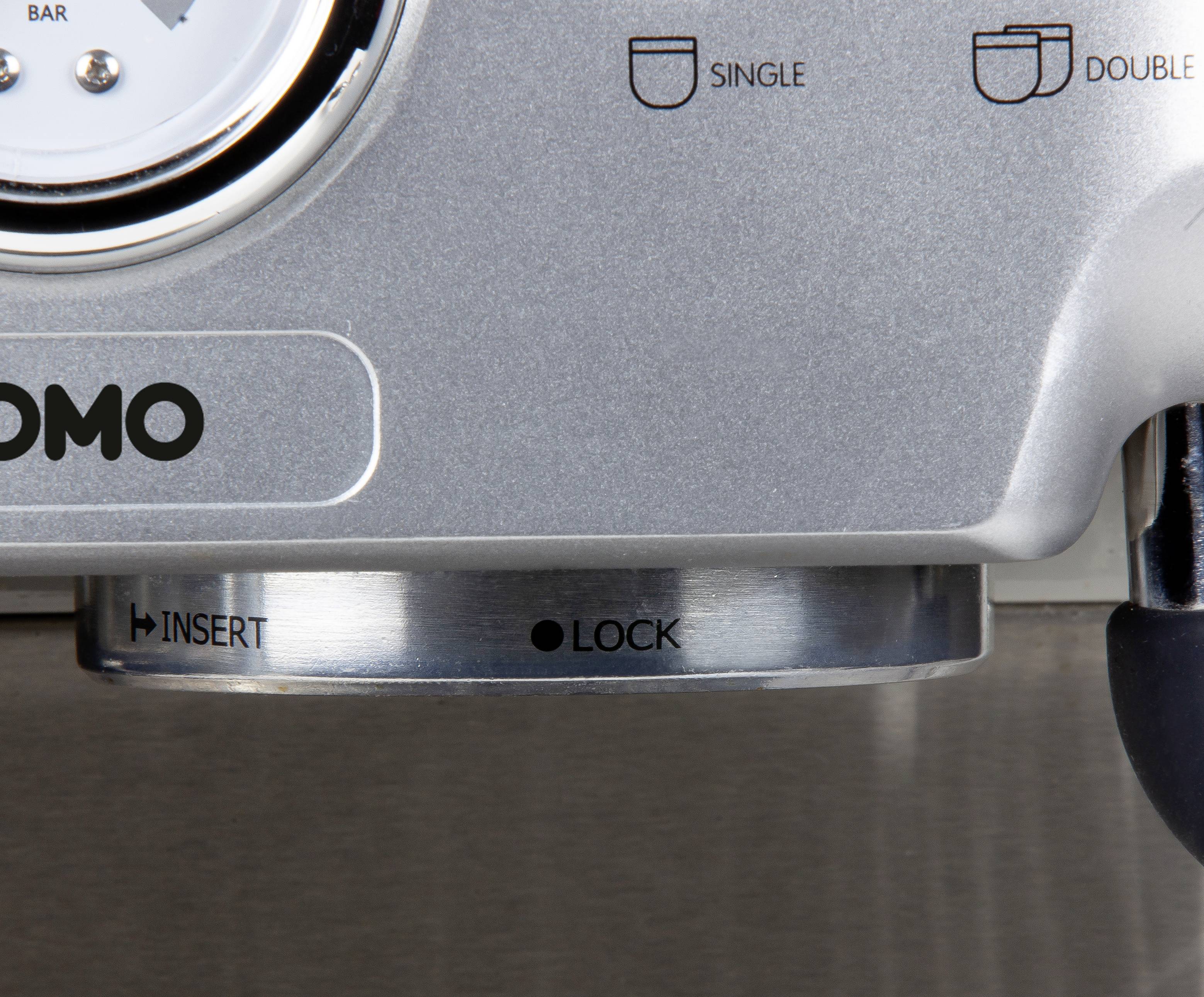 'Metallteil einer Kaffeemaschine mit Drehknopf. Markierungen zeigen 'Insert' und 'Lock'. Anzeige für Bar-Druck sichtbar.'