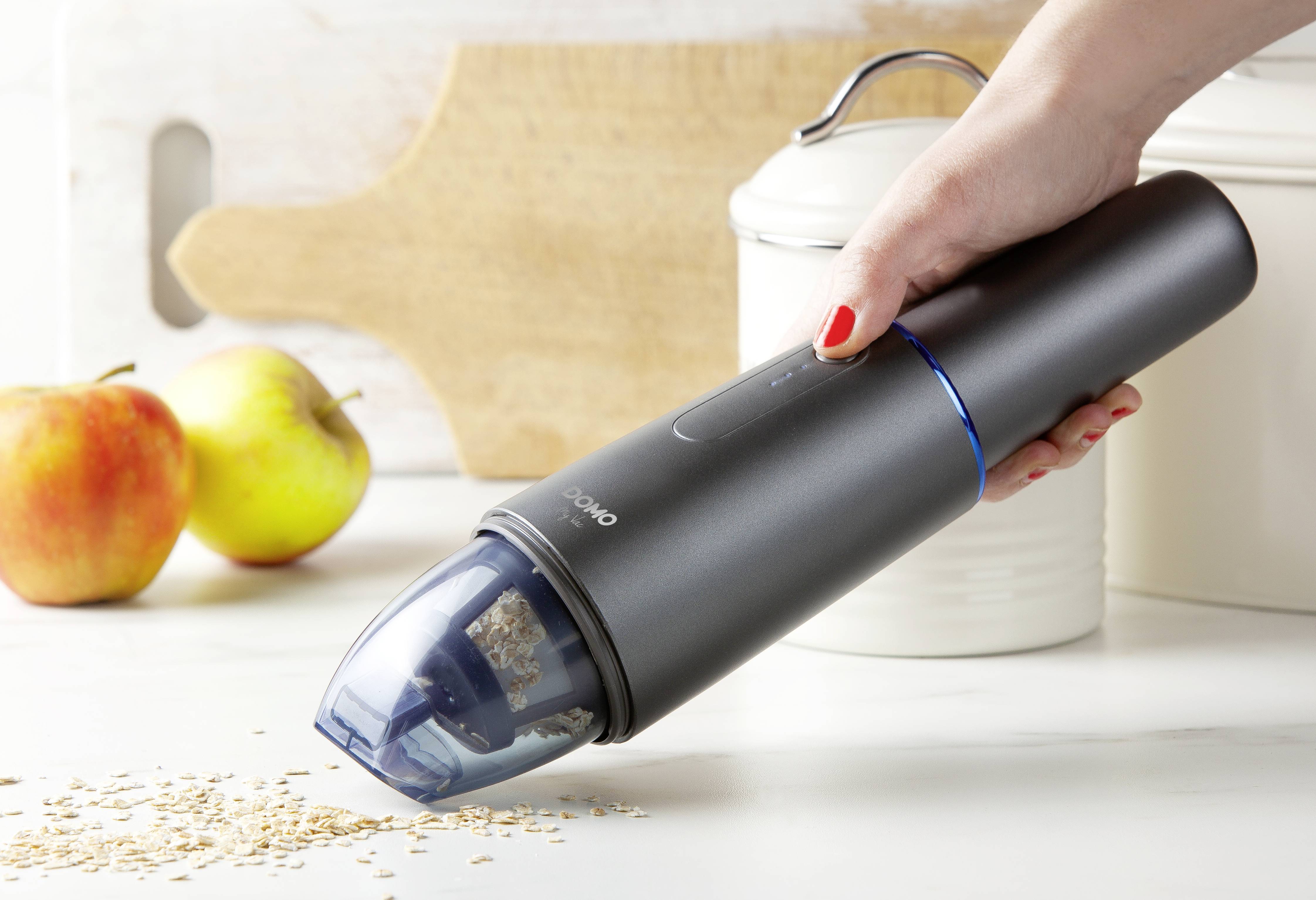 Eine Hand hält einen tragbaren Staubsauger, der auf einer Küche Arbeitsplatte verstreute Körner aufsaugt. Im Hintergrund sind zwei Äpfel.