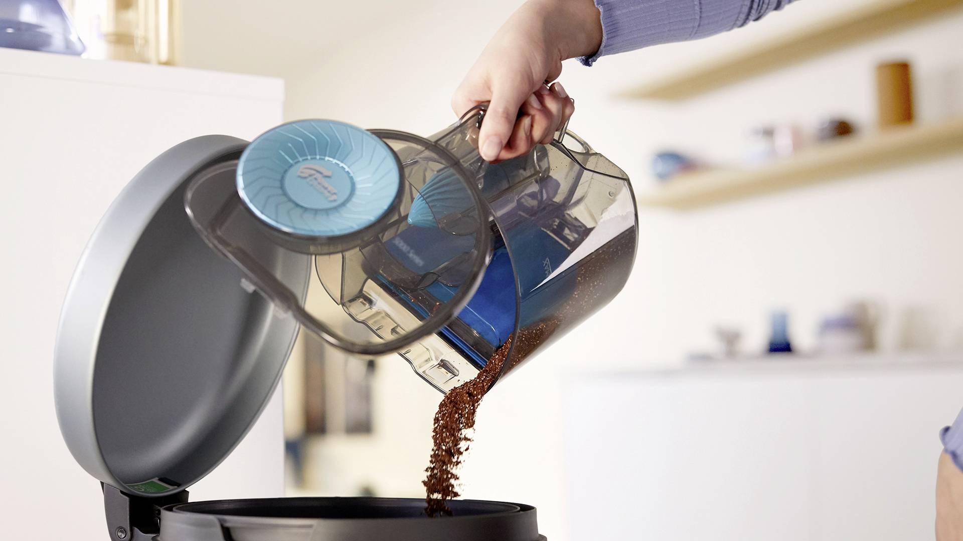 Eine Person leert den Staubbehälter eines beutellosen Staubsaugers in einen Mülleimer.