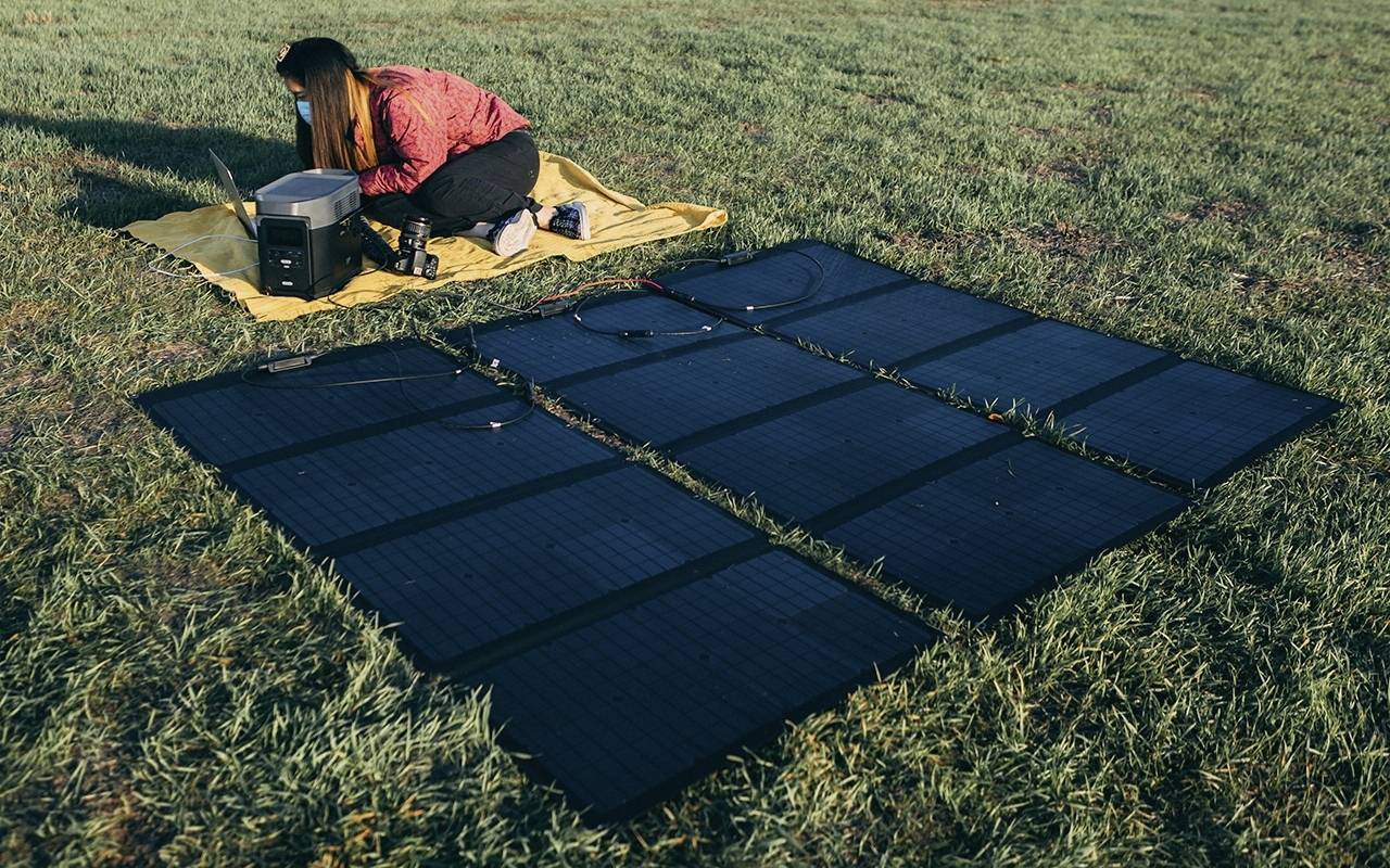Eine Person sitzt auf einer gelben Decke im Gras und arbeitet mit einem Laptop. Vor ihr sind Solarmodule ausgebreitet.